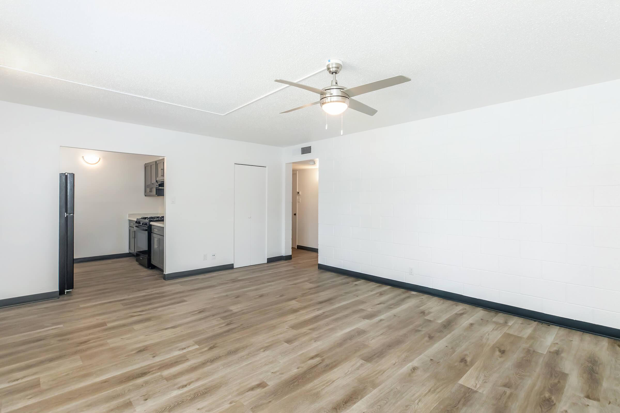 A spacious living room with new laminate flooring, featuring a ceiling fan and light fixture. To the left, a doorway leads to a kitchen area with stainless steel appliances. The walls are painted white, and the overall ambiance is bright and modern.