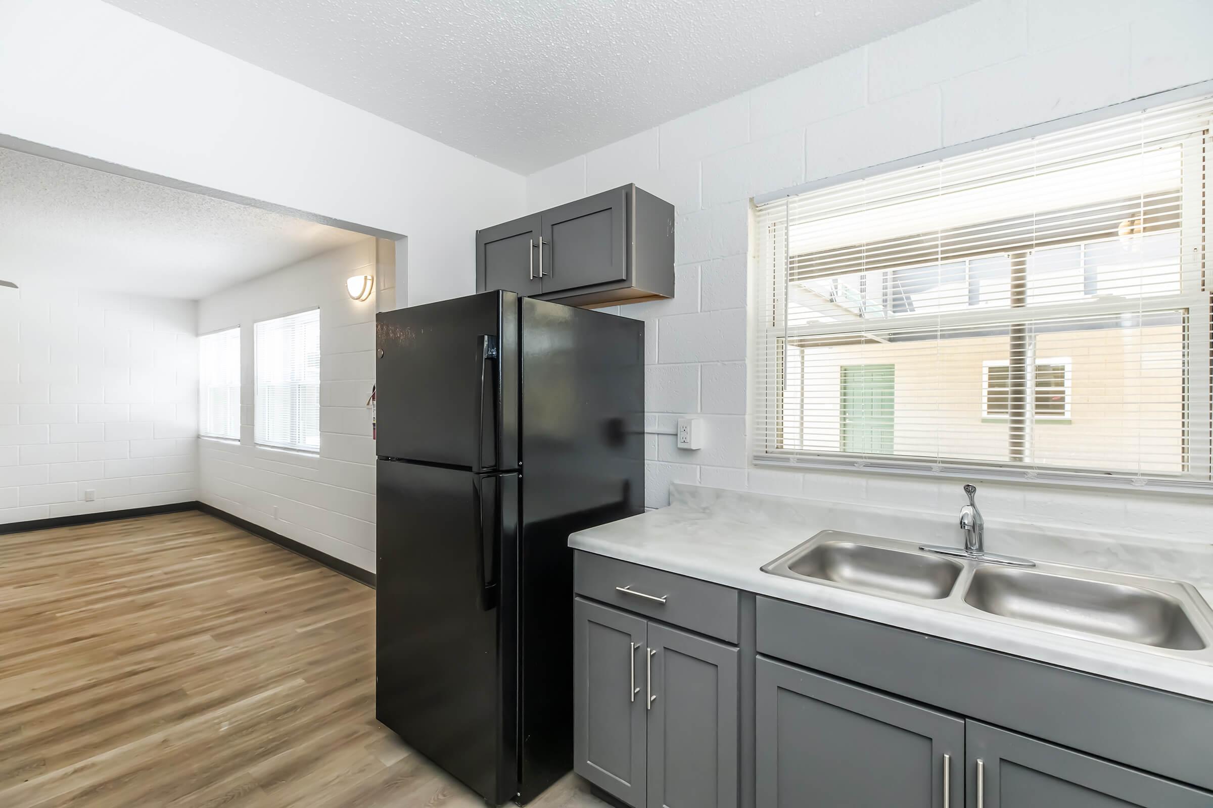 A modern kitchen featuring a black refrigerator, gray cabinetry, and a double sink. Light hardwood flooring extends into a connected living area with large windows allowing natural light. The walls are painted white, creating a bright and open atmosphere.