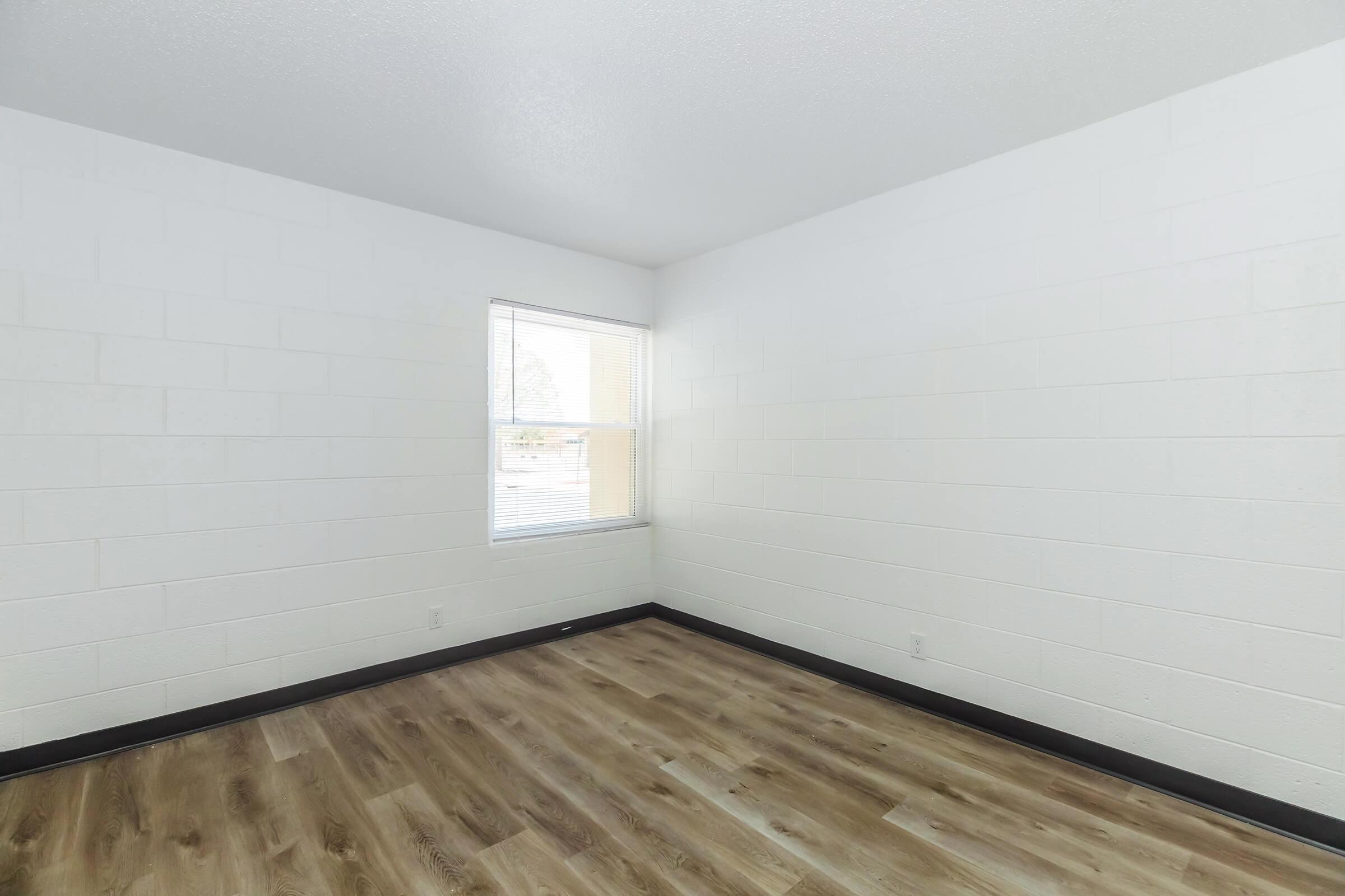 A vacant room with white block walls and a single window letting in natural light. The floor is covered in light brown wood-like laminate, and there are no furnishings or decorations, creating a spacious and minimalistic appearance.