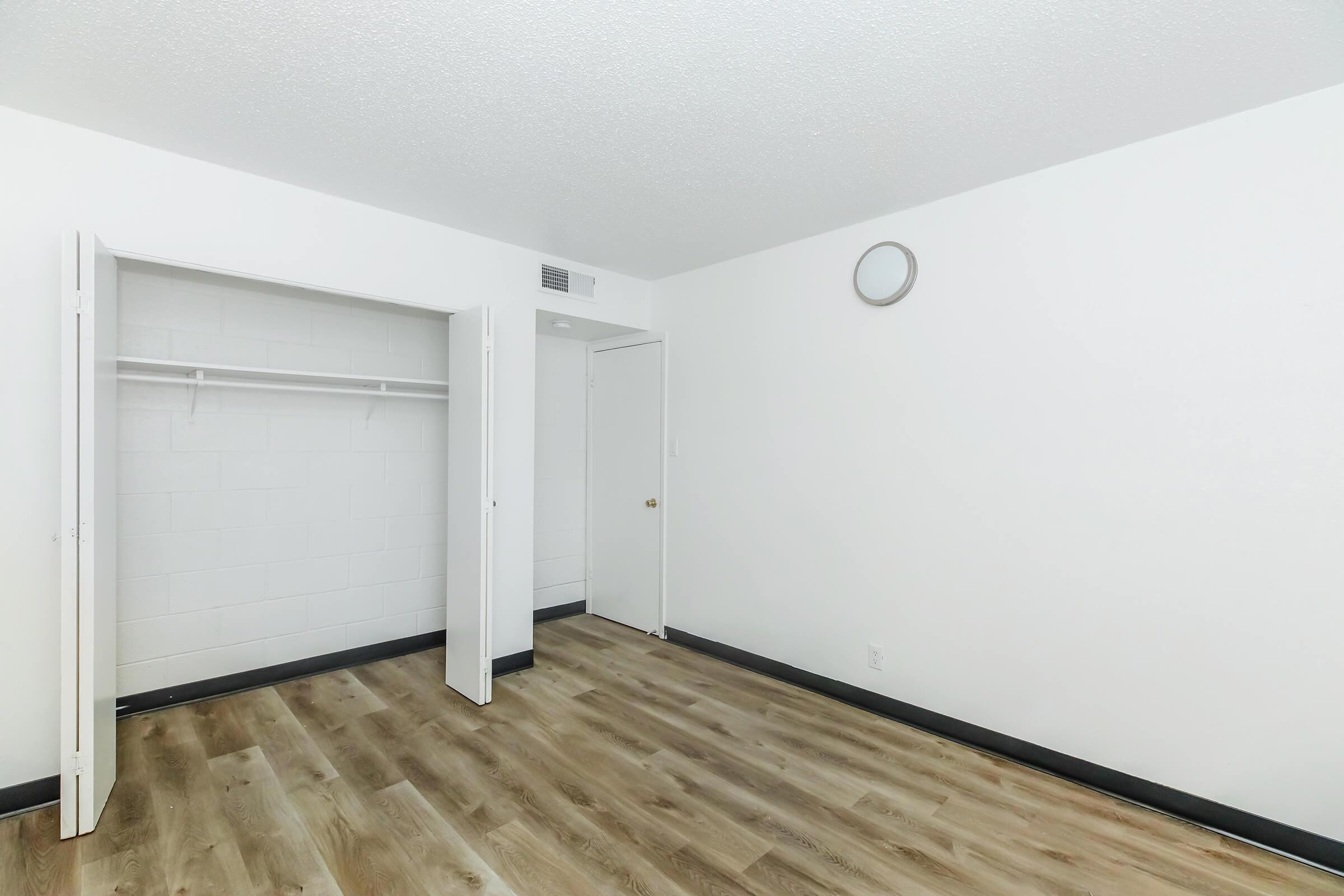 A minimalistic room featuring light-colored walls and a wooden floor. It has two open closet doors that reveal a hanging rod. A small, round clock is mounted on the wall. The space is bright and empty, suggesting it is ready for furnishing. A door leads to another area.