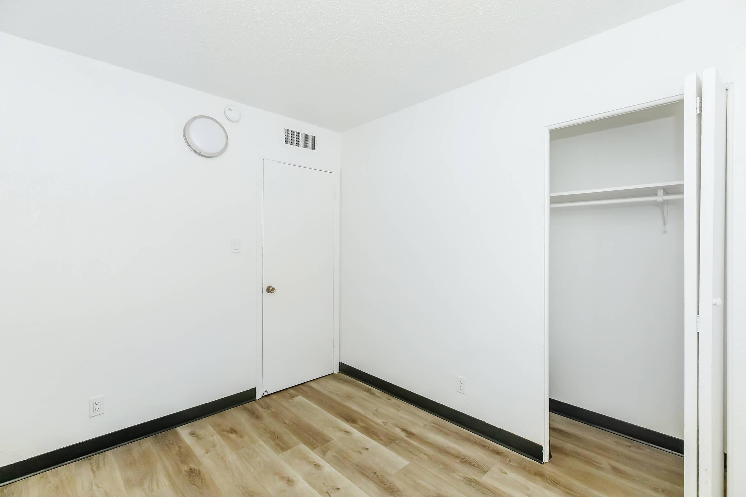Empty room with white walls, a door on the right, and an open closet. The floor features light-colored wood laminate. A small round light fixture is mounted on the ceiling. The space is unadorned and minimalistic, suggesting potential for personalization or use as a bedroom or storage area.