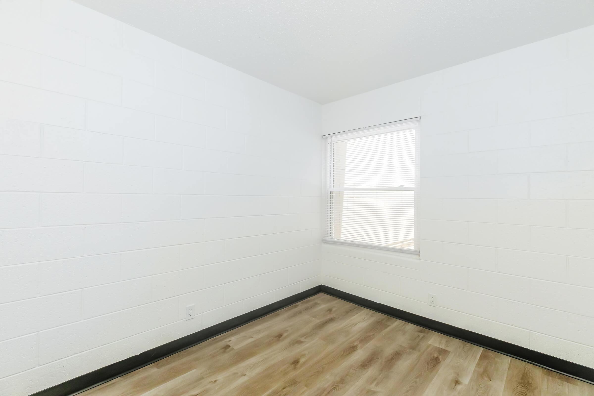 Empty room with white cinder block walls, a single window with blinds, and light-colored wooden flooring. The space is well-lit and has a minimalist design, providing a sense of openness and simplicity.