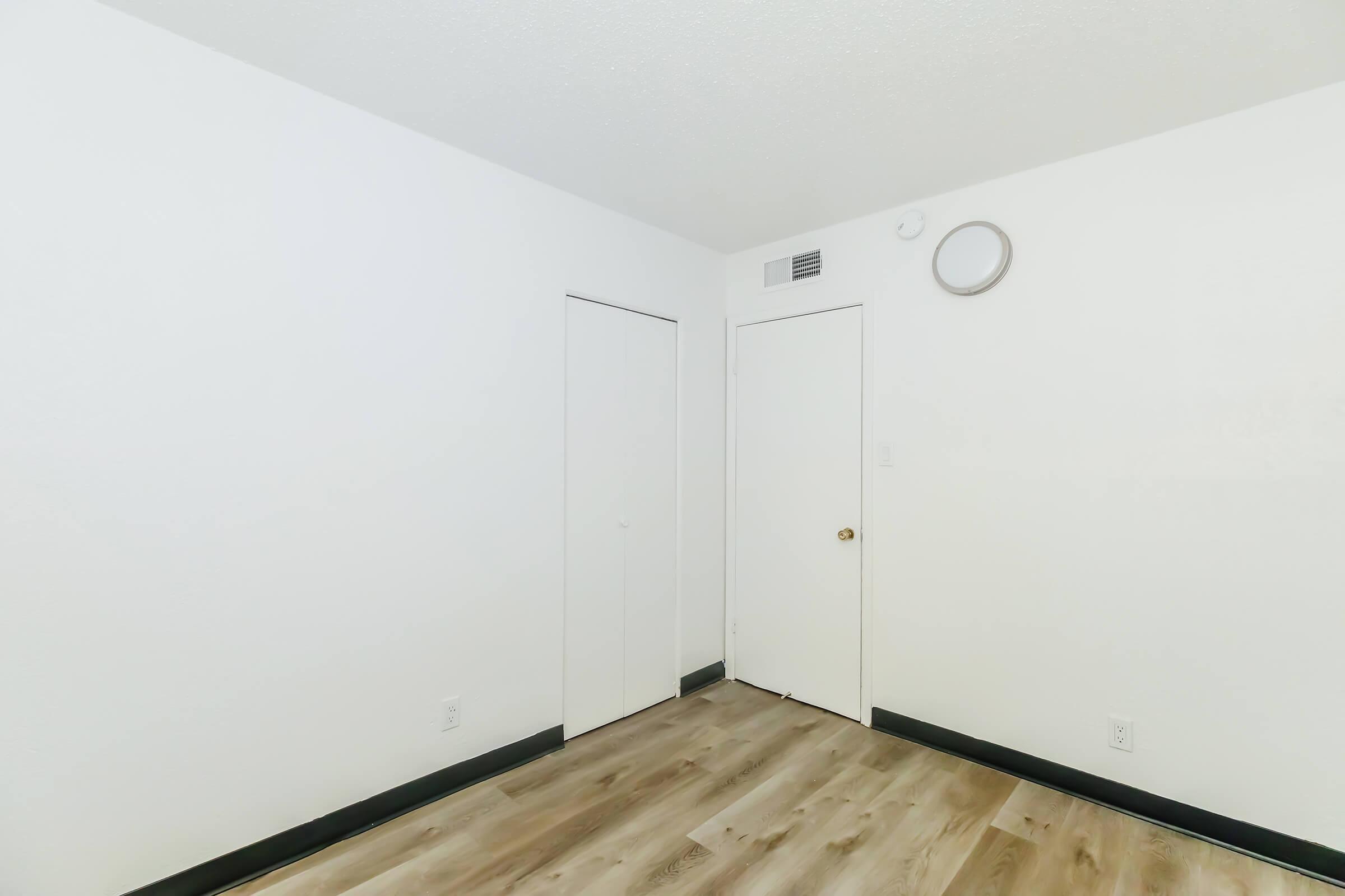A small, empty room with white walls and a wooden floor. There's a closed closet door on one side and a closed main door on the opposite wall. A circular wall-mounted fixture is present near the ceiling. The room appears minimalist and lightly lit.