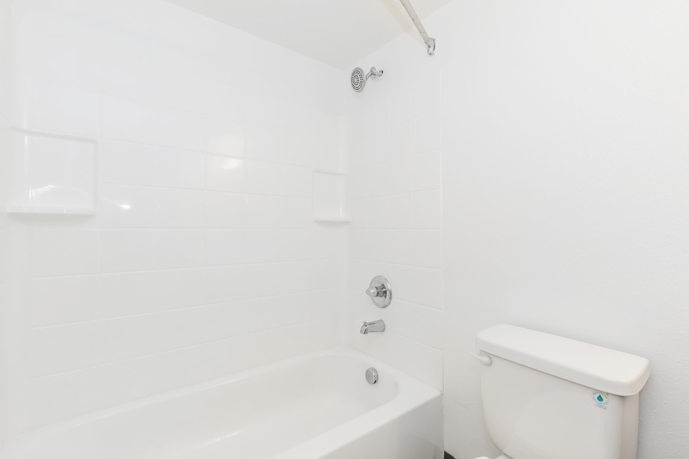 A clean, minimalistic bathroom featuring a white bathtub with a showerhead, white tiled walls, and a white toilet. The overall decor is simple and modern, contributing to a bright and airy atmosphere.