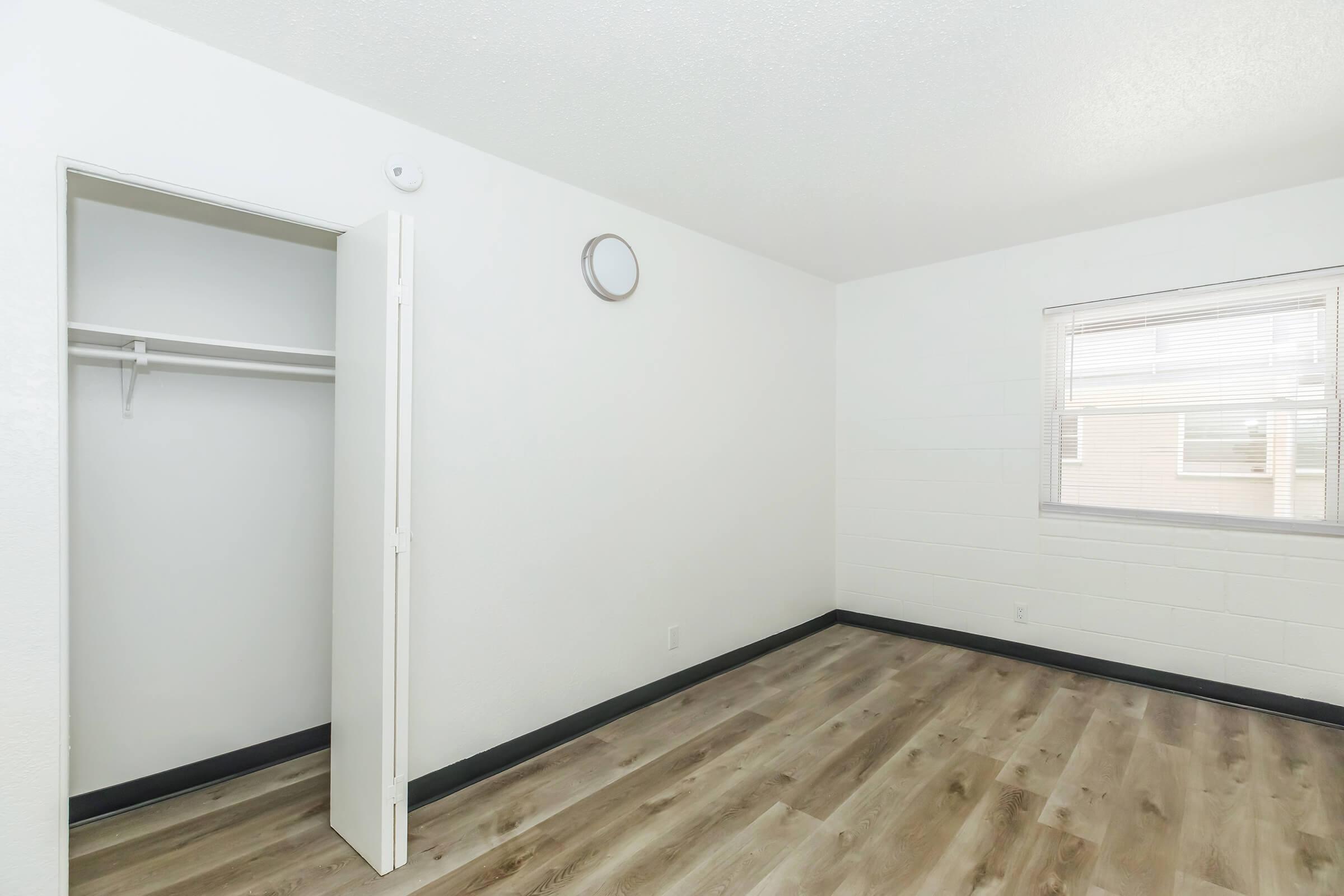 A vacant room featuring light-colored walls, a window with blinds, and wooden flooring. There is an open closet with a hanging rod, a round wall clock, and minimal decor, creating a simple and spacious atmosphere.