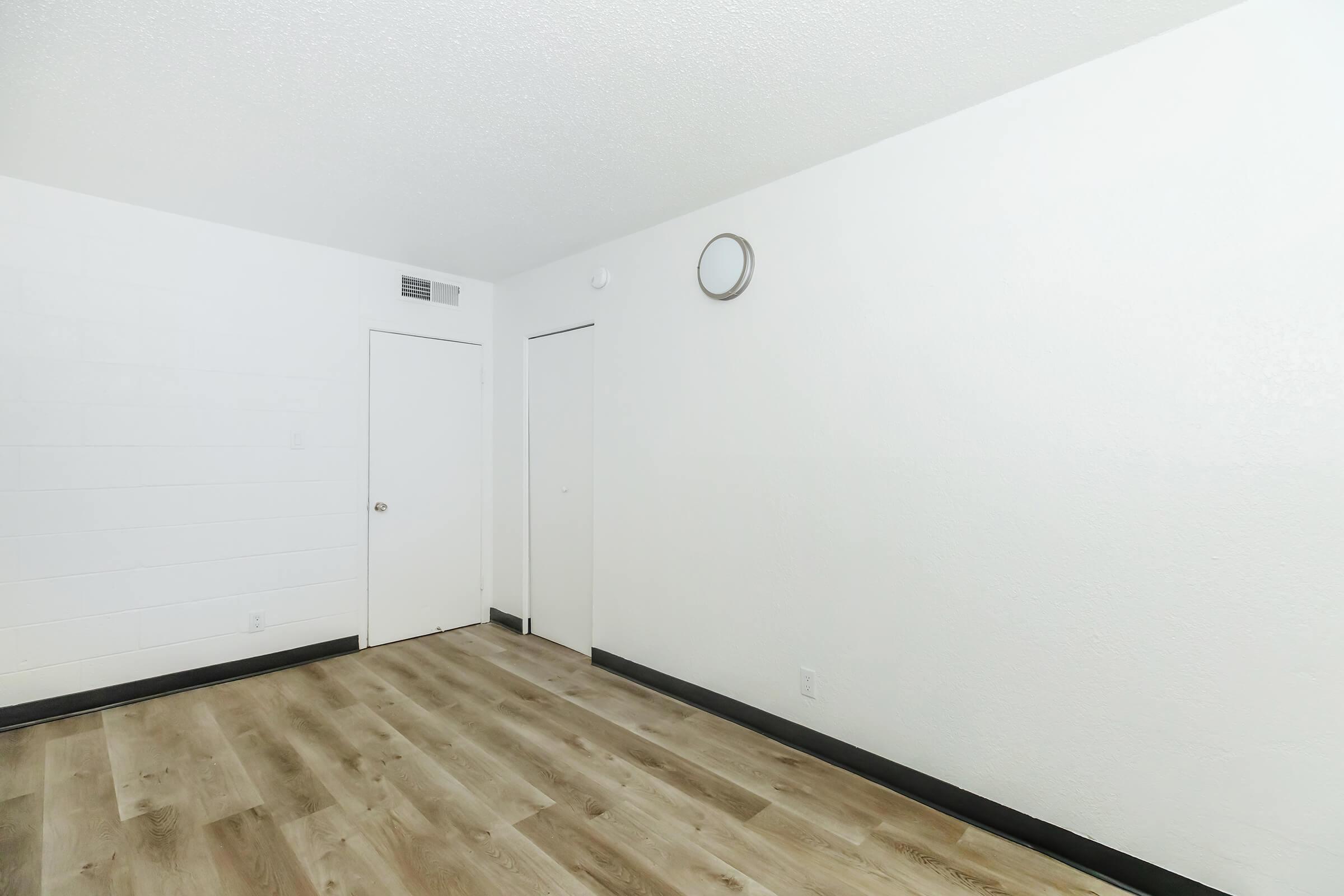 Empty room with white walls, light wooden flooring, and a small round wall clock. There are two doors, one leading to a closet and the other an entryway, both closed. The room is well-lit and has a minimalistic design, creating a spacious and clean appearance.
