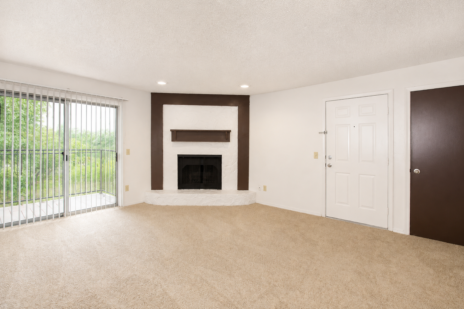 A spacious living room featuring beige carpeting, a white stone fireplace with a dark wooden mantel, and natural light coming through sliding glass doors leading to an outdoor area. The room includes a white door and a dark brown door, creating a cozy and welcoming atmosphere.