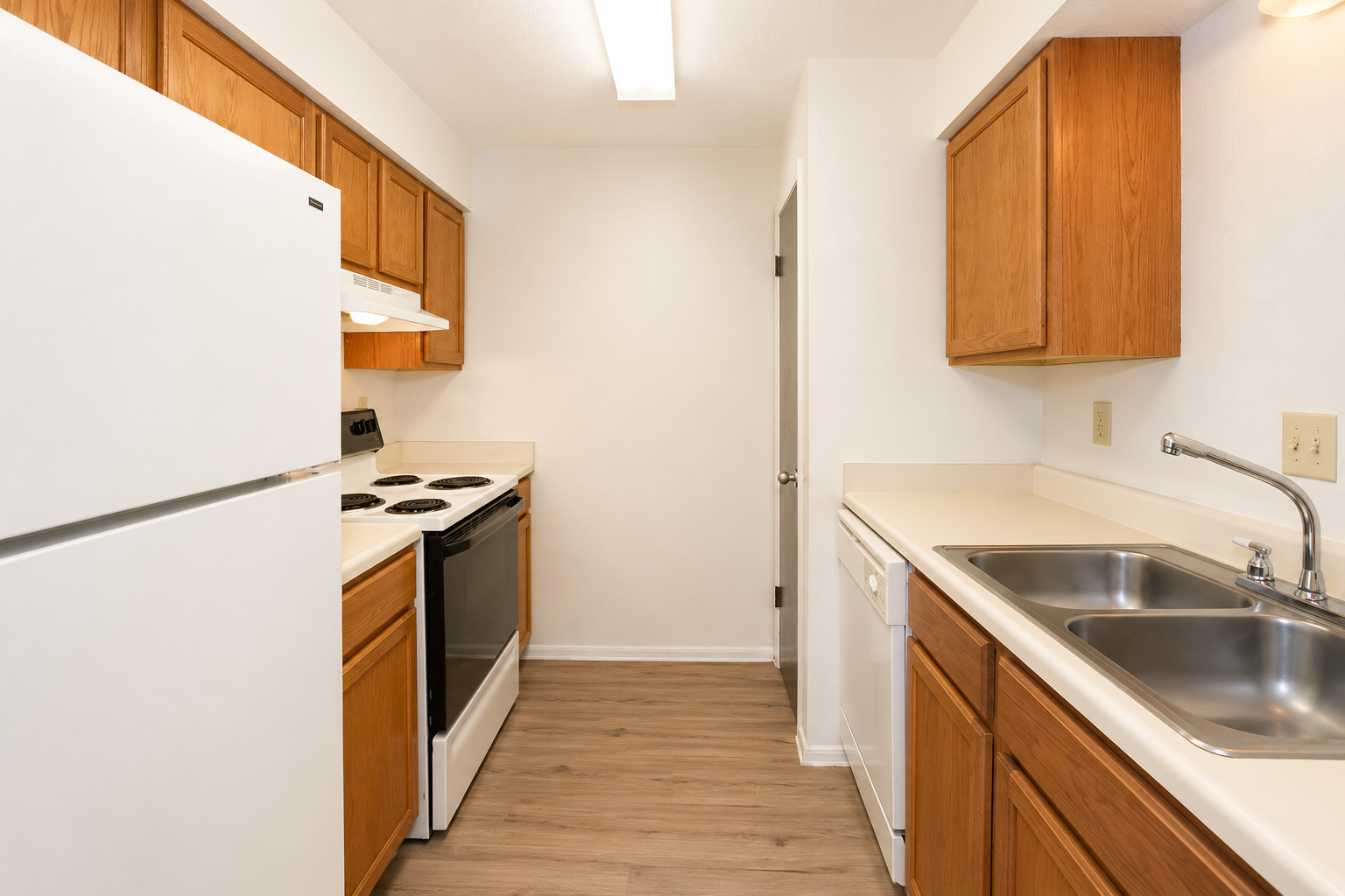 A modern kitchen featuring wooden cabinets, a white refrigerator, an electric stove with an overhead microwave, and a double sink. The countertops are light-colored, and the flooring is a wood-like laminate. The kitchen is well-lit with overhead lighting and has a clean, organized appearance.