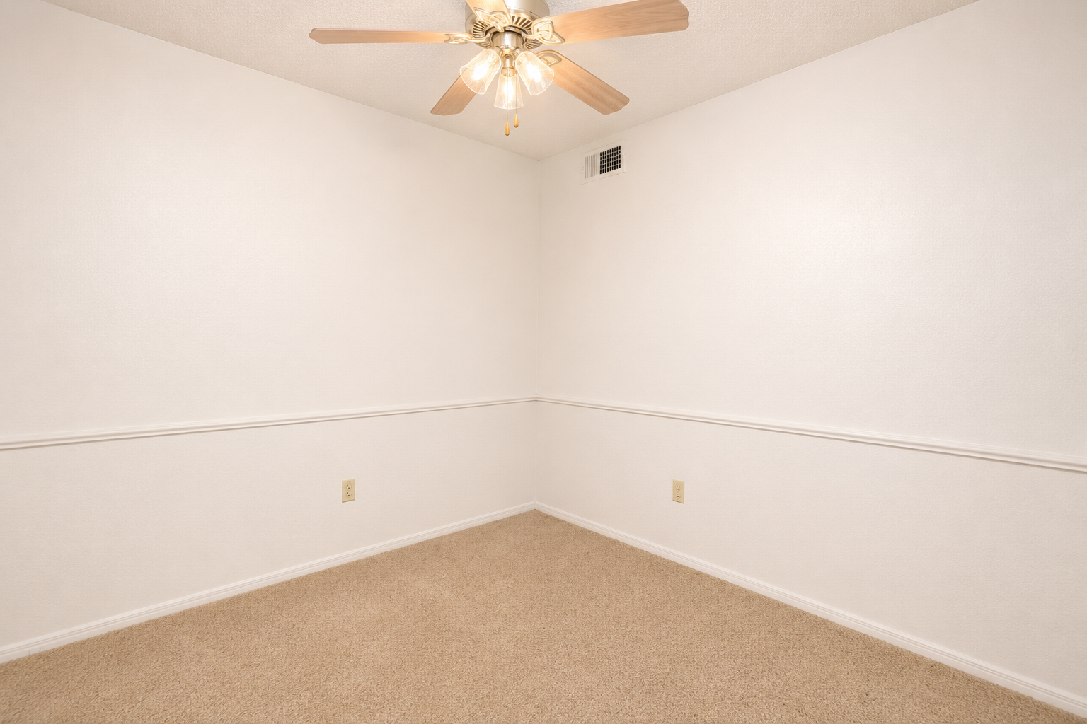 A spacious, empty room with light beige carpet and white textured walls. The ceiling features a ceiling fan with wooden blades, and there is a white chair rail running along the wall. The room has no furniture or decorations, creating a simple and open atmosphere.