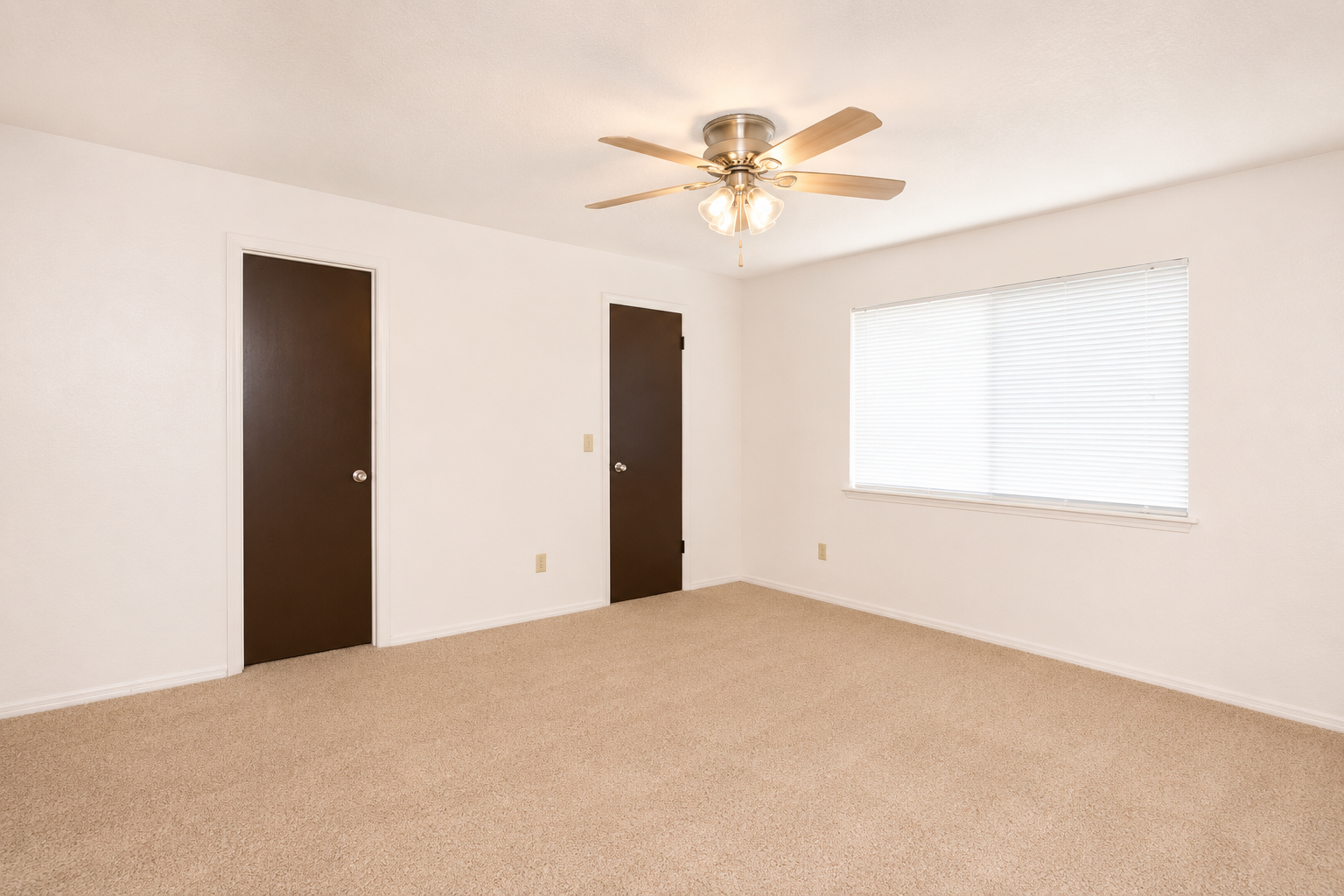 A clean and empty room featuring light beige carpet, white walls, and a ceiling fan with a light fixture. There are two closed dark brown wooden doors and a window with horizontal blinds allowing natural light to enter. The overall atmosphere is bright and spacious, suitable for various uses.