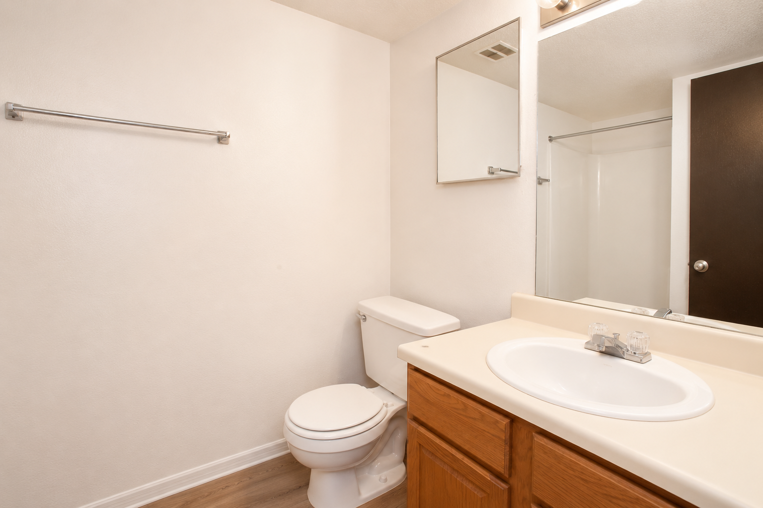 A clean and simple bathroom featuring a white toilet, a sink with a light wood vanity, a large mirror, and a shower area. The walls are light-colored, and the flooring is a wooden style. A towel rack is mounted on the wall opposite the sink.
