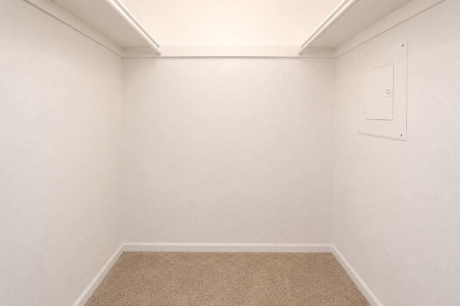 An empty, well-lit closet with white walls and a beige carpet. There are two horizontal shelves near the ceiling and a small electrical panel on one wall. The overall appearance is clean and minimalist, conveying a sense of spaciousness.
