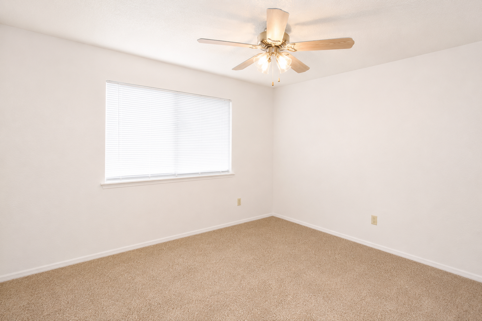 Empty room featuring a light-colored carpet, a ceiling fan with a light fixture, and a window with closed blinds. The walls are painted white, creating a bright and airy atmosphere, and there are no furnishings present, giving the space a clean and minimalist appearance.