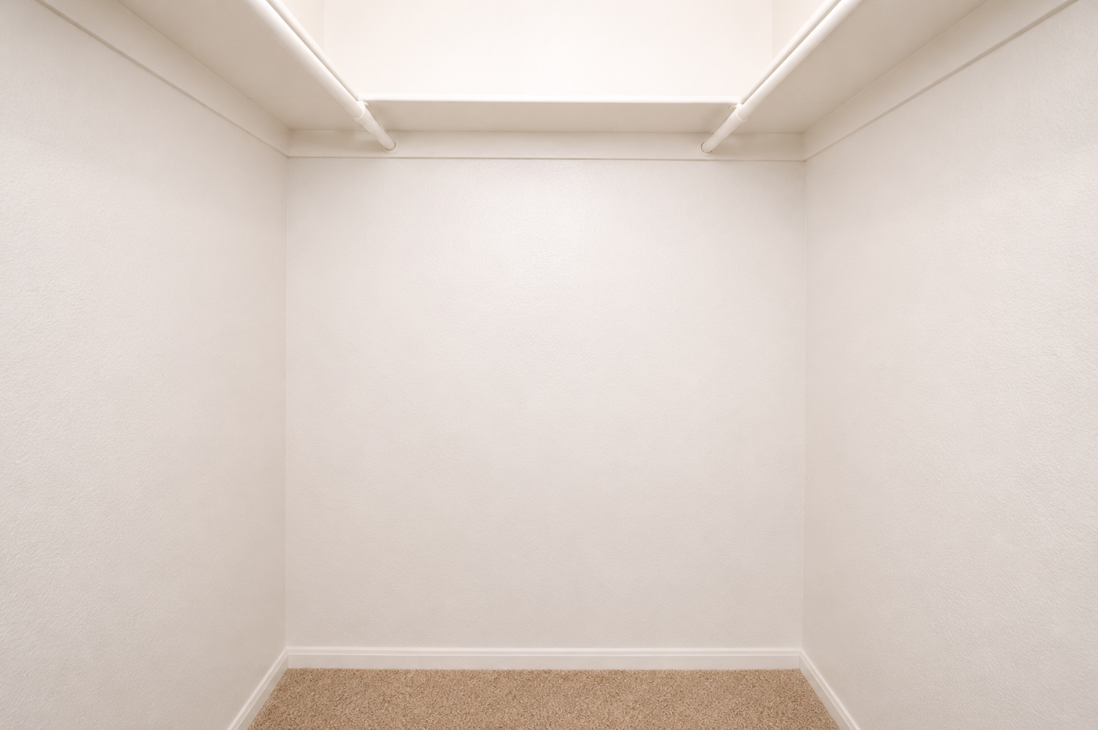 A spacious, empty closet with white walls and a carpeted floor. The closet features two shelves above, providing potential storage space. The overall feel is clean and minimalistic, with no items or decorations present.