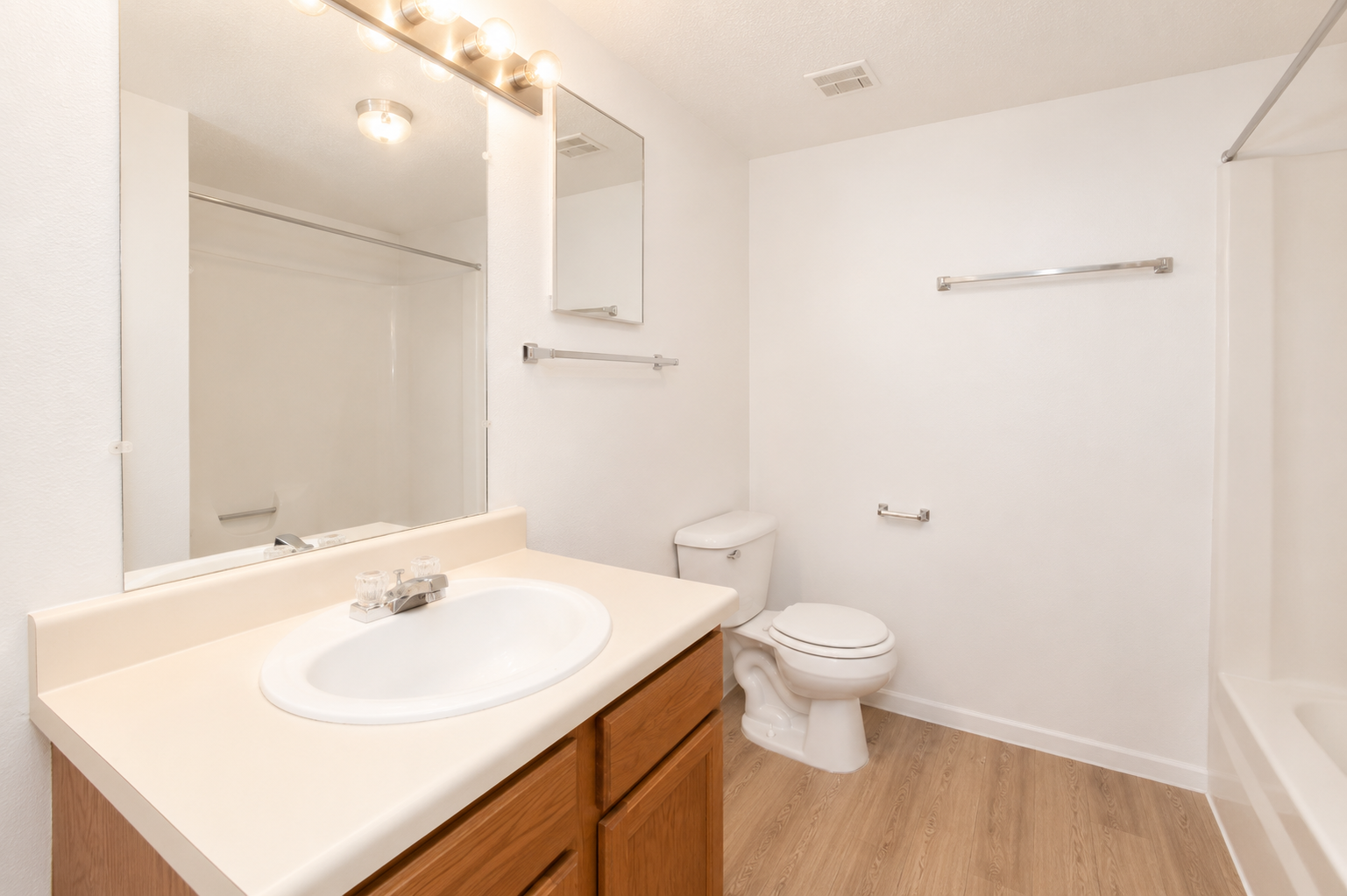 A clean and bright bathroom featuring a white sink with a single faucet, wooden cabinetry, a wall-mounted mirror, a toilet, and a shower area with a glass door. The walls are painted light, and the floor has a wood-like finish, creating a spacious and modern look.