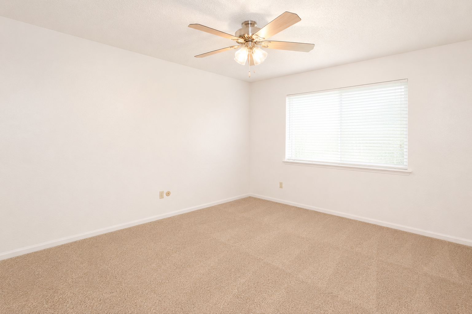 A spacious, empty room with a light beige carpet and off-white walls. There is a ceiling fan with wooden blades and a single window covered by white blinds, letting in natural light. The room is devoid of furniture, creating a clean and simple aesthetic.