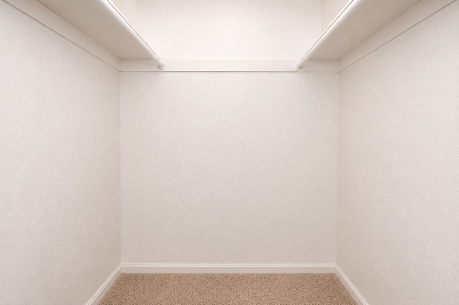 Empty walk-in closet with white walls and a light beige carpet. There are two shelves fixed to the upper wall, creating a clean and minimalistic look. The space is unoccupied, providing a fresh canvas for organizing or storing items.