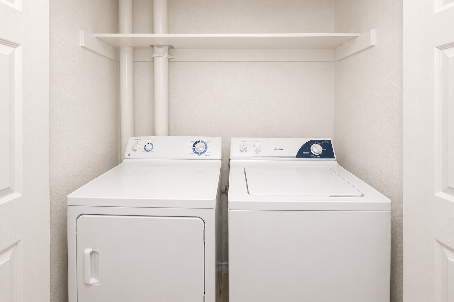 A small laundry room featuring a white washing machine and a white dryer side by side. The walls are painted a light color, and there is a shelf above the appliances. Pipes are visible on the wall behind the machines, and the space is well-lit, creating a clean and organized appearance.