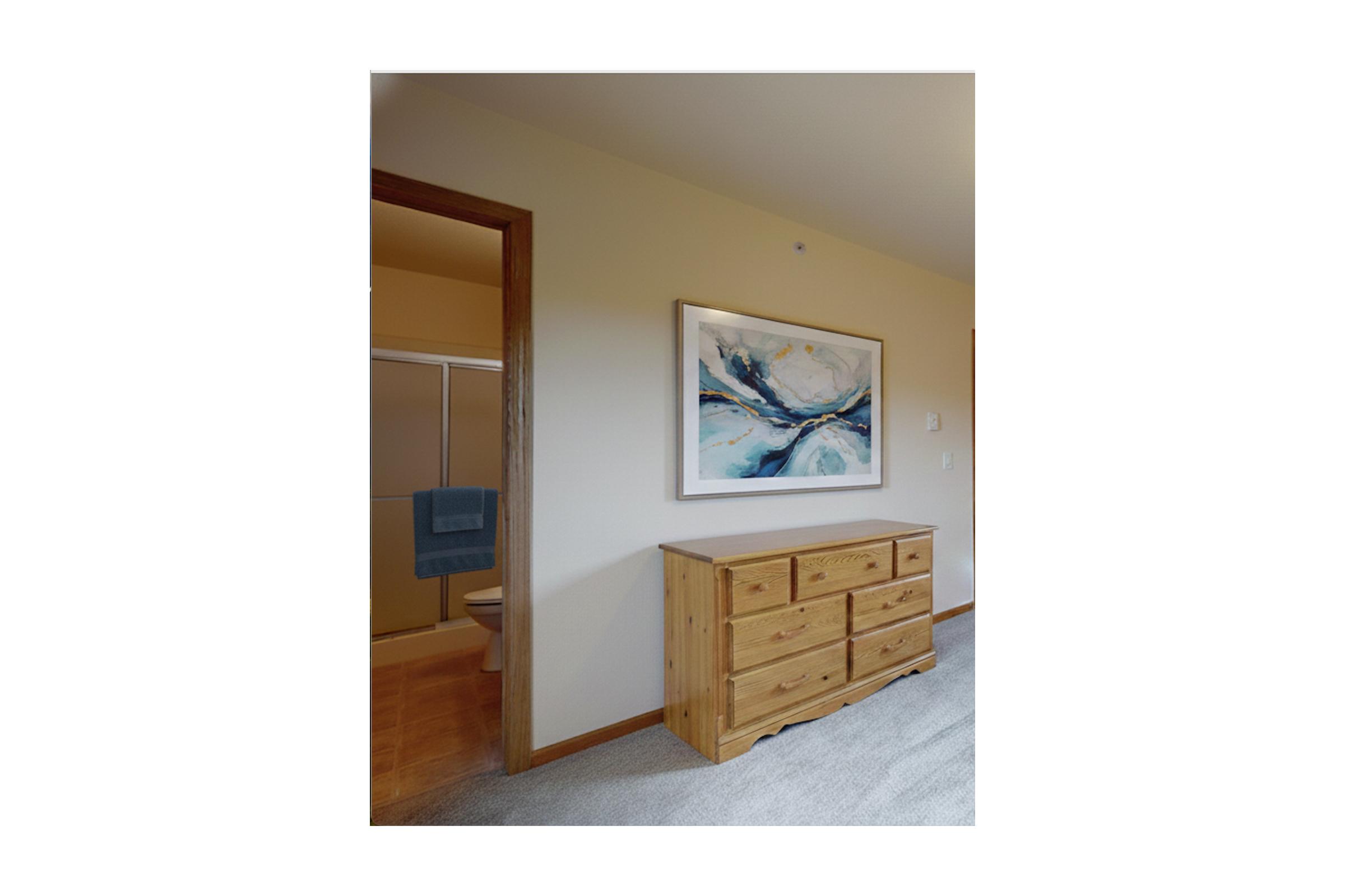 A cozy interior scene featuring a wooden dresser with multiple drawers against a wall. Above the dresser hangs a large abstract painting with blue and white tones. A doorway leads to a bathroom on the left, partially visible, with a towel hanging on a rack. The room has neutral-colored walls and carpet.