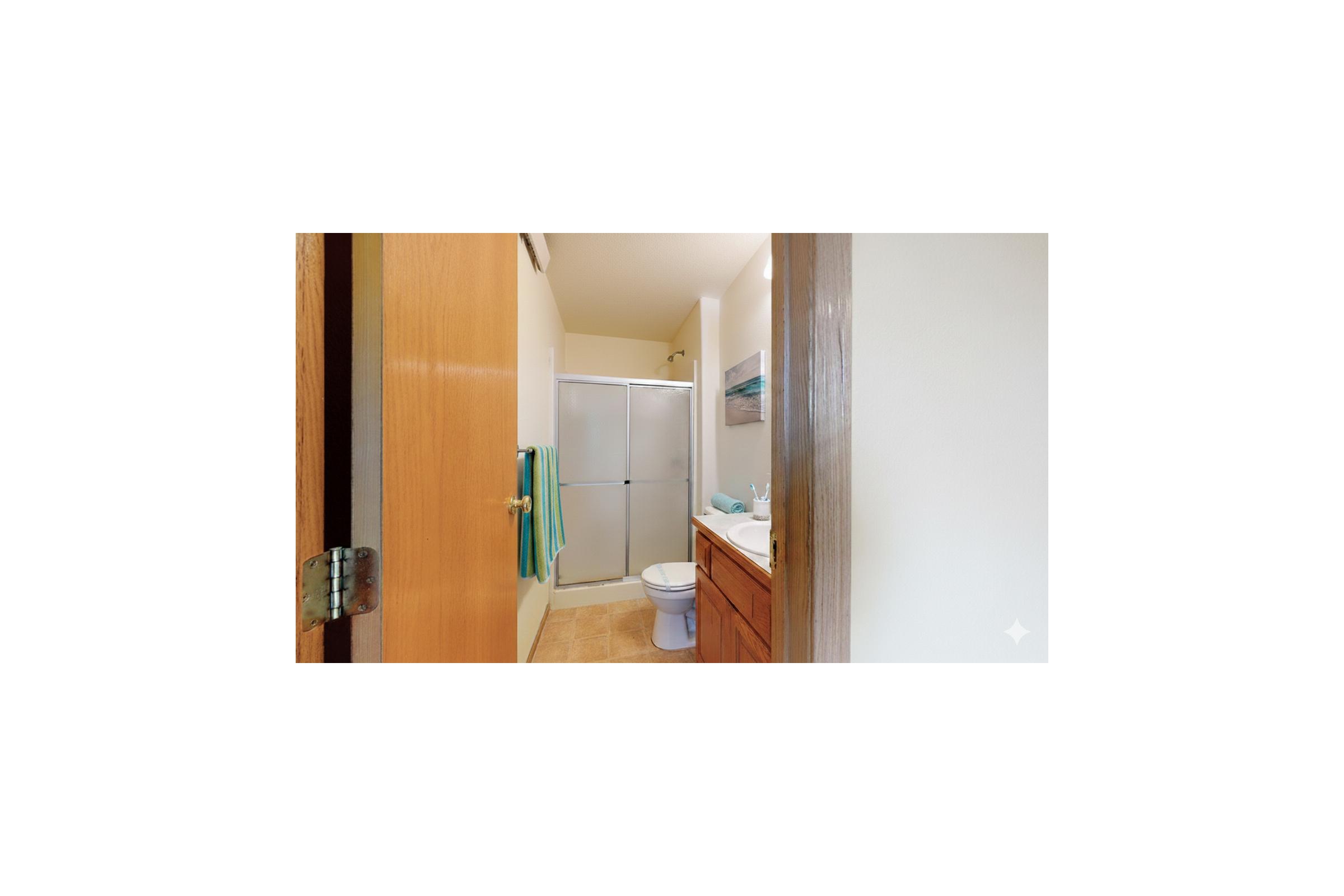 View of a bathroom featuring a shower area with sliding doors, a toilet, and a sink with a mirror above it. The room has light-colored walls and wooden cabinetry, with a blue towel hanging near the shower. The entrance is partially visible from a wooden door.