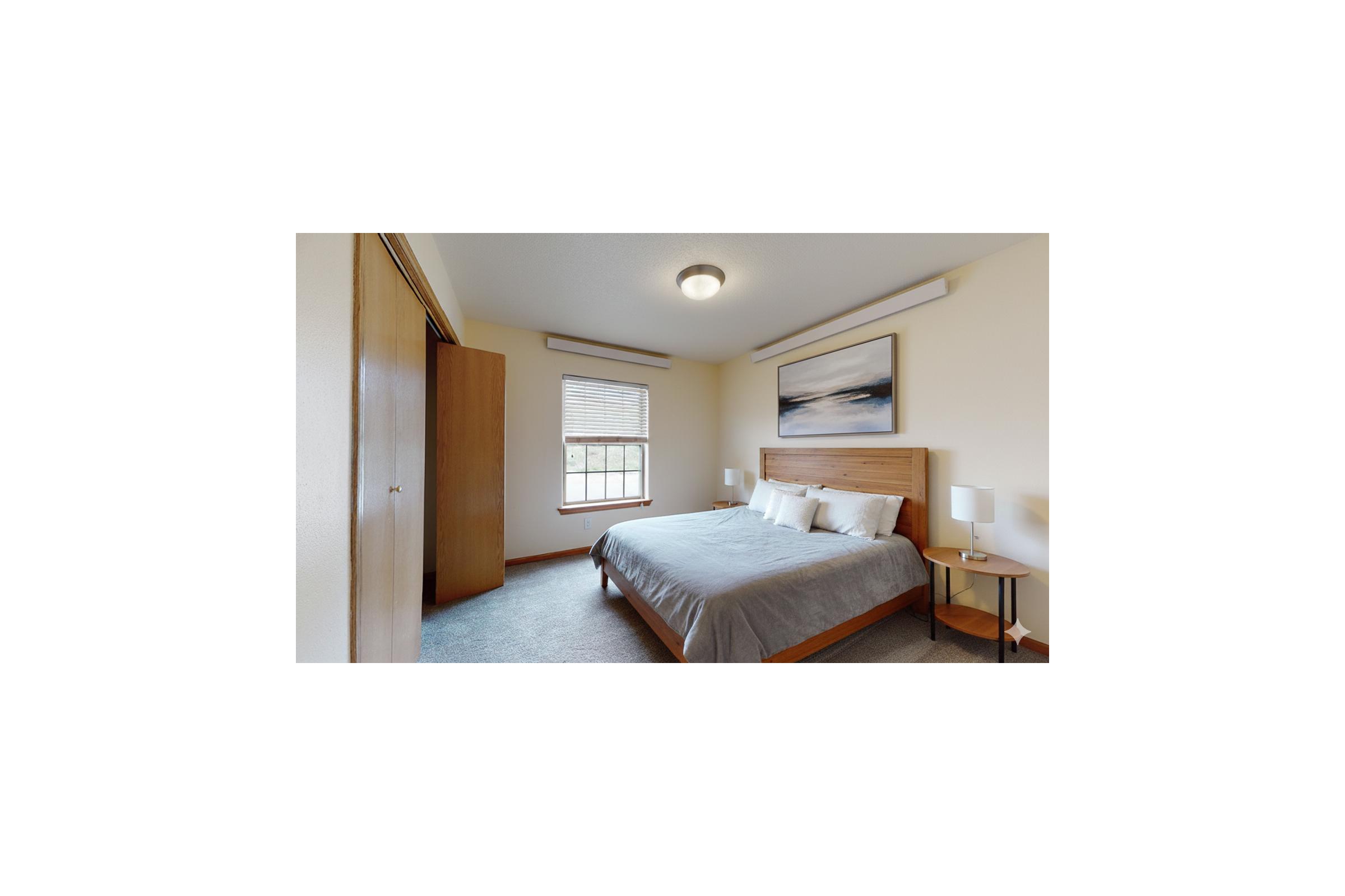 A cozy bedroom featuring a queen-sized bed with a light gray comforter, two bedside tables with lamps, and a wooden wardrobe. A large window lets in natural light, and a piece of abstract art hangs above the bed, creating a serene atmosphere. The walls are painted in a soft neutral color.