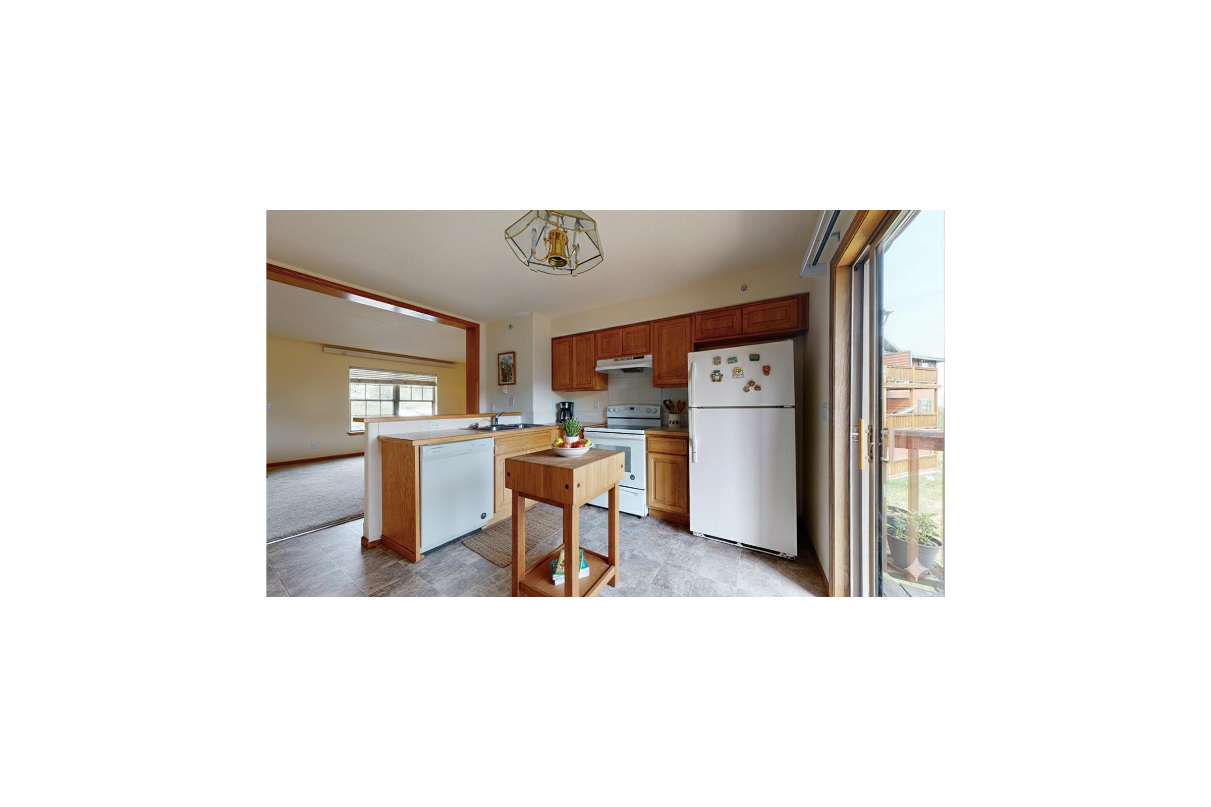 A modern kitchen featuring wooden cabinets, a white refrigerator, and a small wooden kitchen island. The space has large windows allowing natural light and a view of the outside. The floor is tiled, and there are minimal decorative items on the counters.