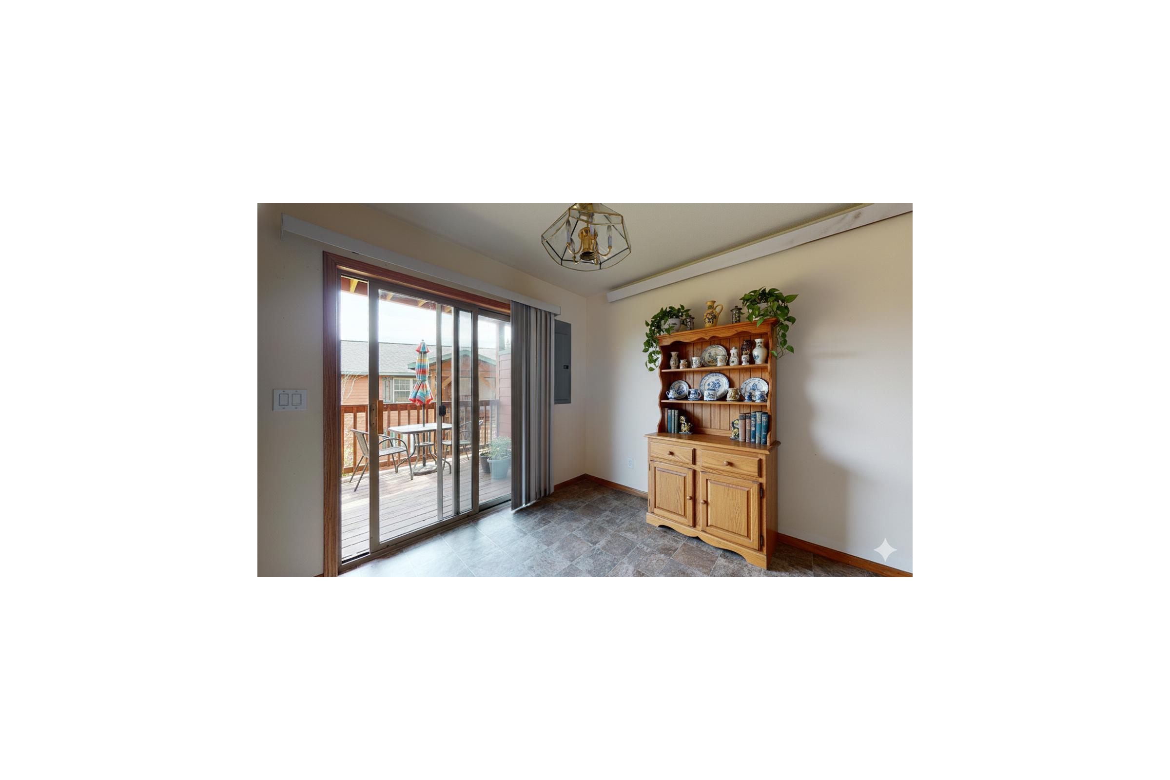 A bright and well-lit room featuring a wooden cabinet filled with decorative items and books. There are sliding glass doors leading to an outdoor deck, with potted plants on either side of the cabinet. The flooring is tiled, adding a cozy touch to the space.