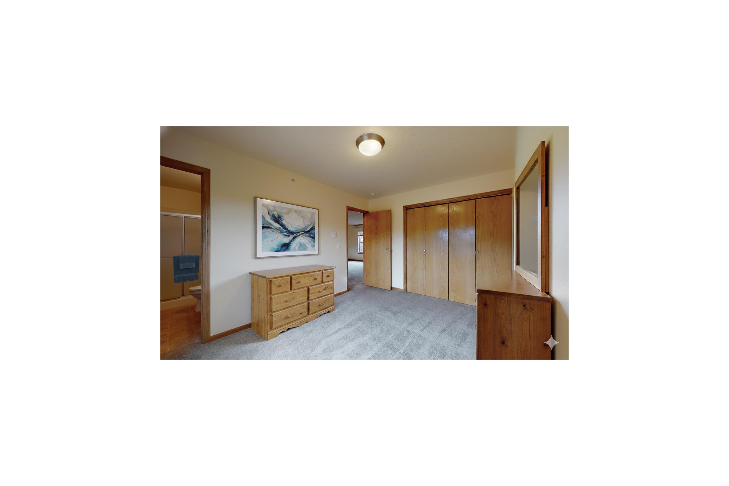 Interior view of a hallway in a home featuring light-colored walls, a carpeted floor, wooden furniture including a dresser and mirrored cabinet, a piece of abstract art on the wall, and doors leading to other rooms. Natural light is present, creating a warm and inviting atmosphere.