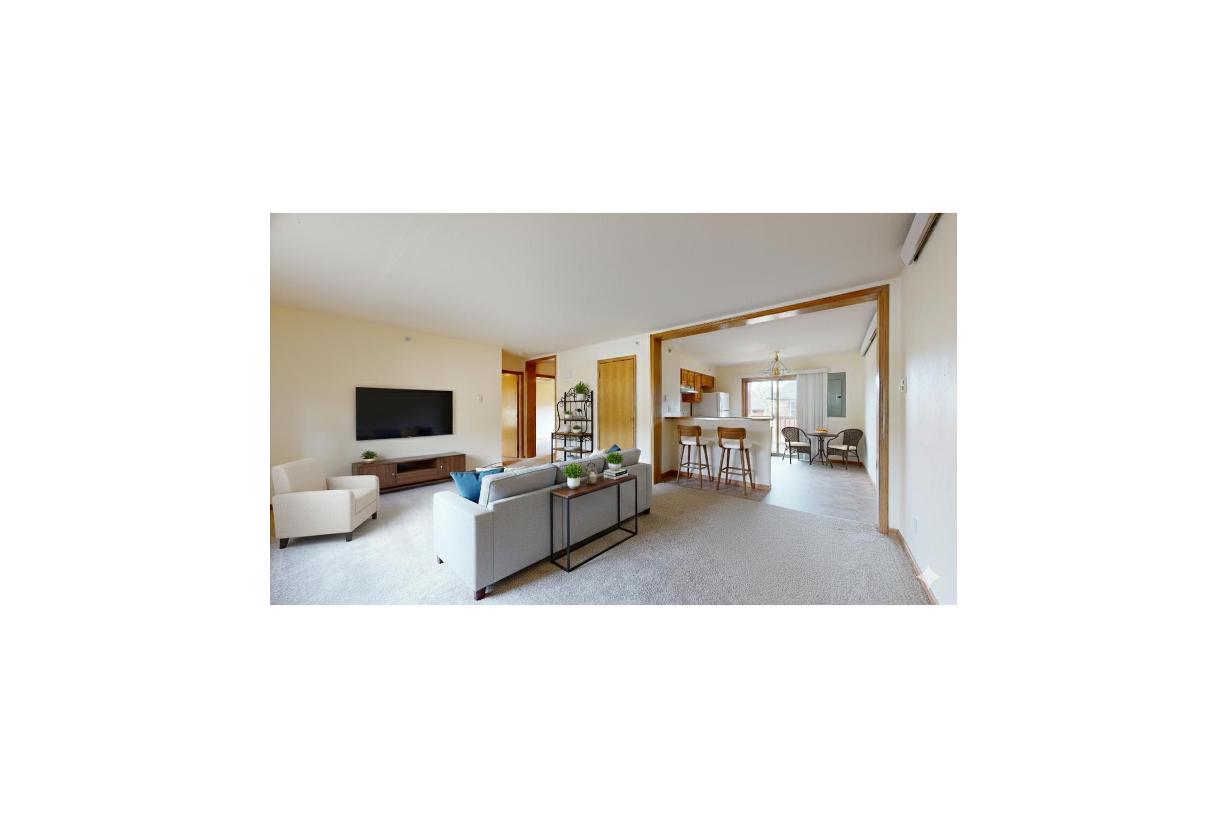 A modern, airy living room featuring a beige armchair, a stylish coffee table, and a large TV mounted on the wall. The space is connected to a dining area with bar stools and a light-colored kitchen in the background. Natural light enhances the open concept design.