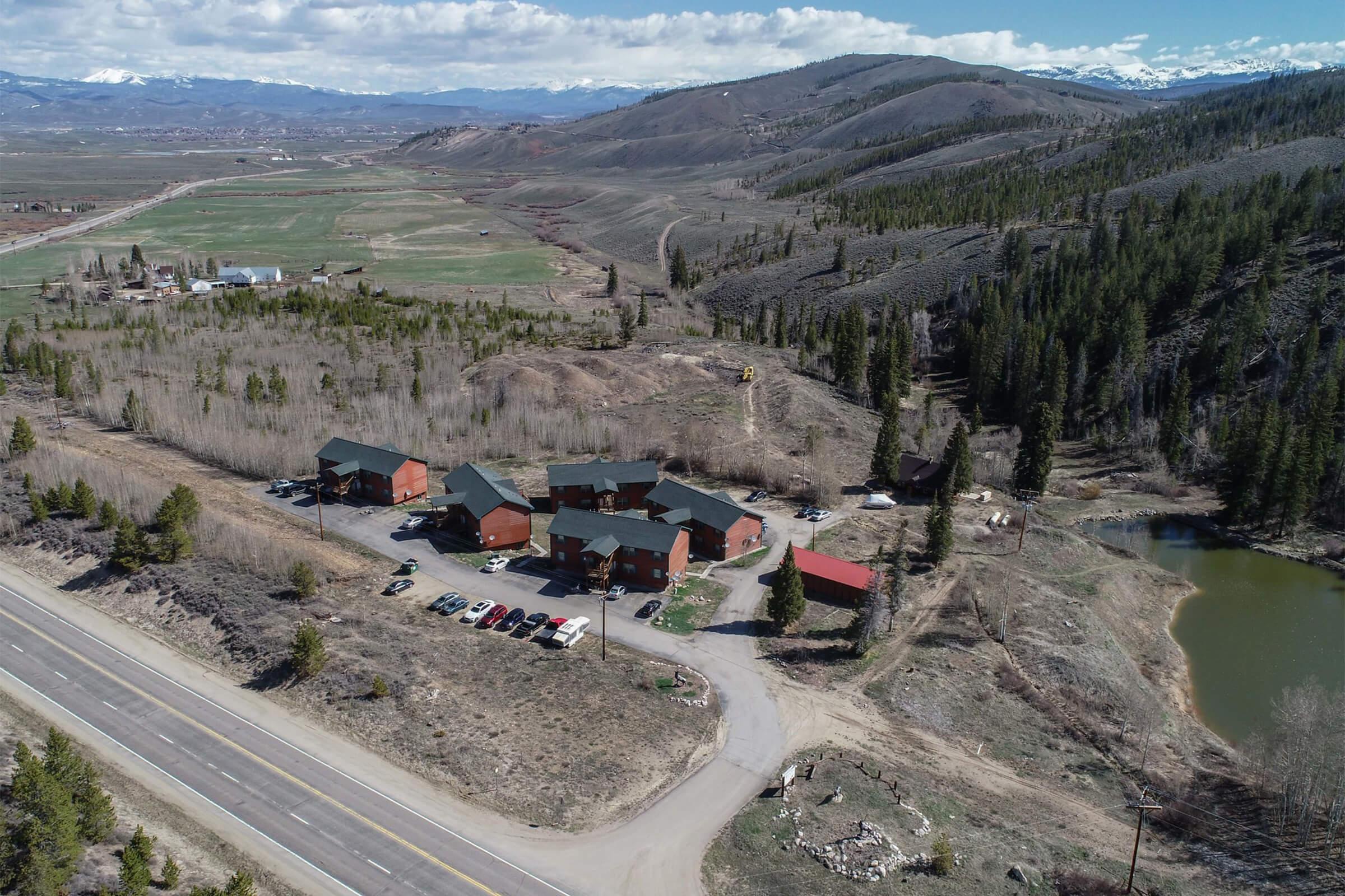 Aerial view of a rural area featuring a cluster of brown buildings with dark roofs, surrounded by trees and mountains. A winding road runs alongside the property, with several parked cars visible. In the background, green fields and distant snowy mountains complete the scenic landscape.