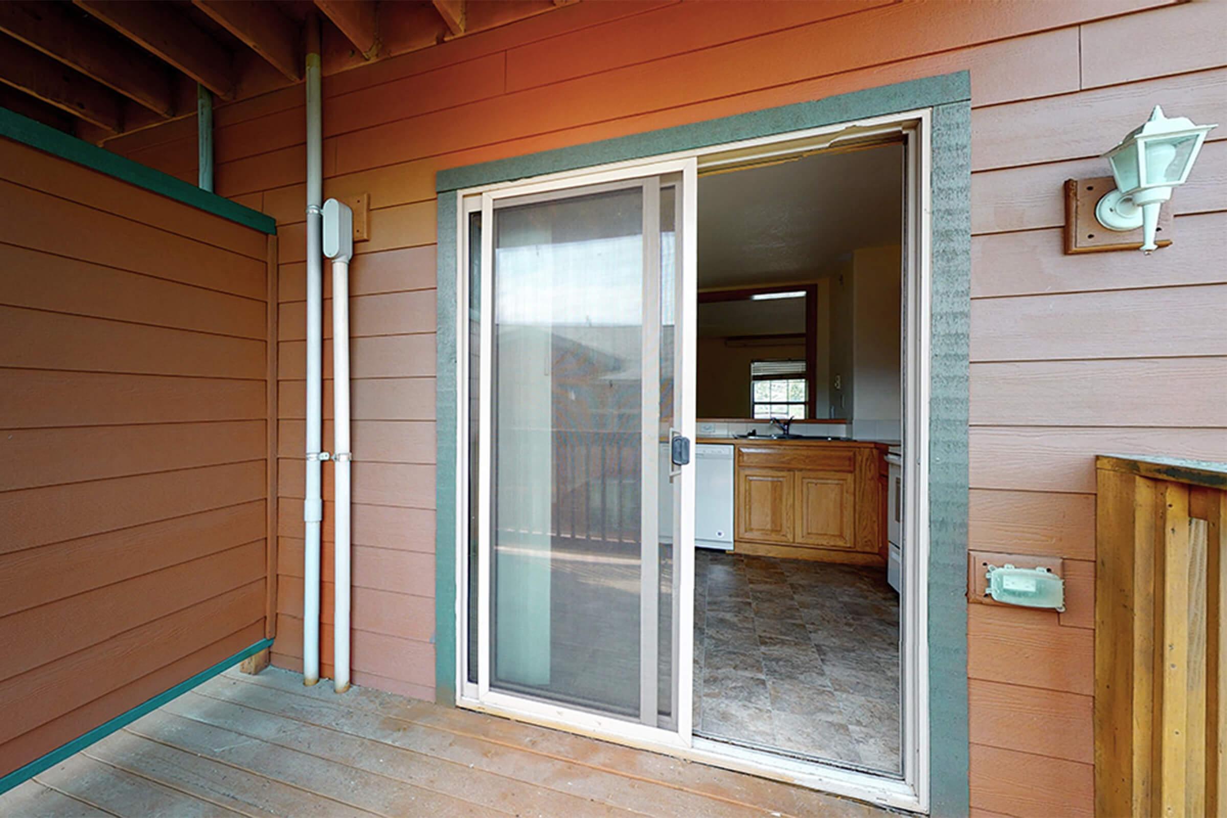 A partially open sliding glass door leads to a kitchen area with wood cabinetry and light-colored walls. The exterior features wooden siding and a light fixture, while the floor is made of tiles. The space appears well-lit and welcoming.