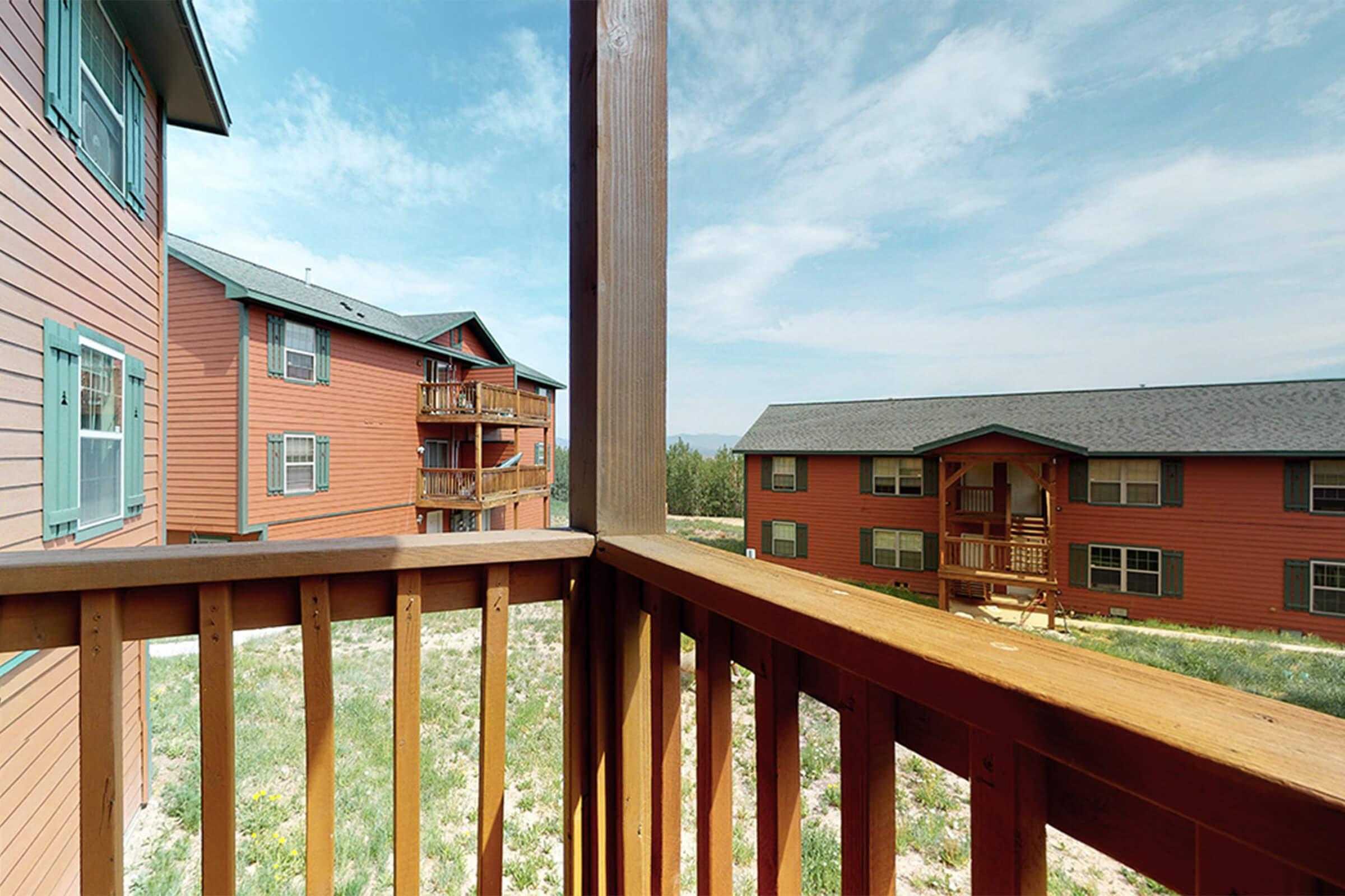 View from a wooden balcony overlooking a community of red-colored apartment buildings. The scene includes green landscaping and a clear blue sky with some clouds, highlighting a tranquil residential environment.