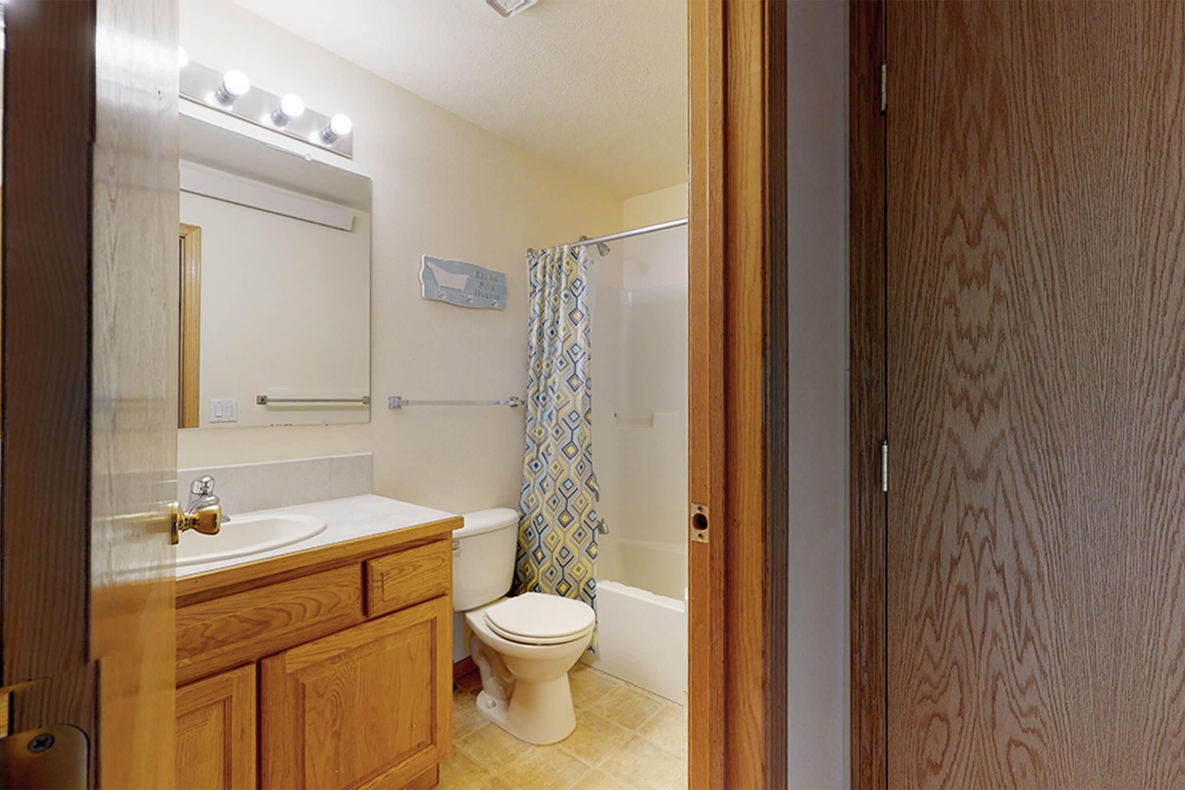 A well-lit bathroom featuring a wooden vanity with a sink, a toilet, and a shower-tub combo. The shower has a decorative curtain with a blue and yellow pattern. Light fixtures are above the mirror, and a door leads to the hallway.