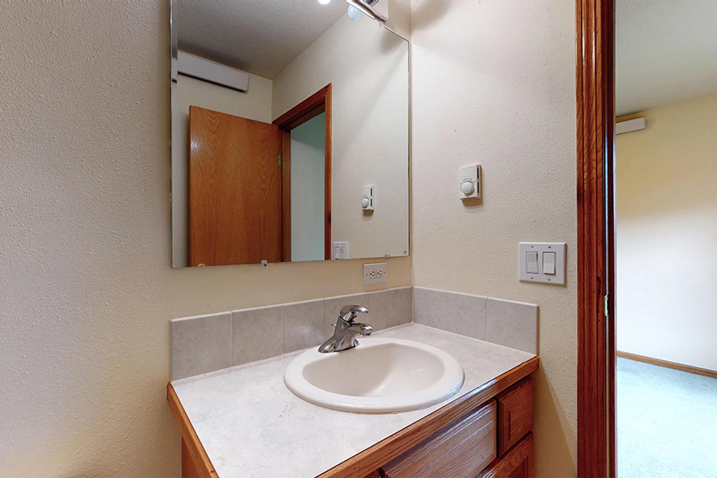 A small bathroom featuring a sink with a single faucet, a mirror above it, and light fixtures. The walls are painted in a light color, and there's a door leading to another room. The floor is tiled, and the countertop is made of light-colored material. The overall space appears clean and well-maintained.
