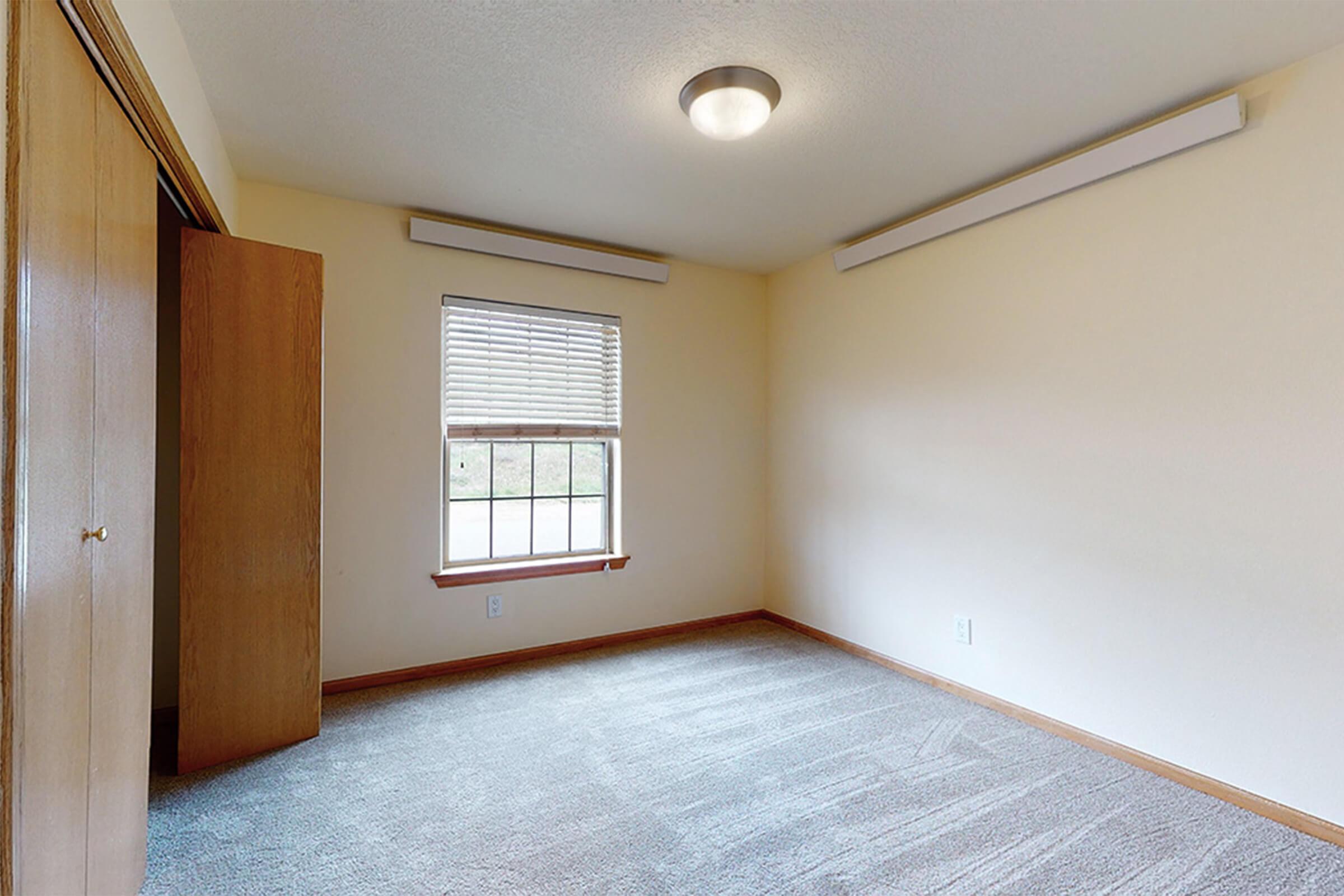A small, vacant room featuring beige walls, a window with blinds, and gray carpet flooring. The room has a ceiling light fixture and two wall-mounted lights. There is a closet with wooden doors on the left side. The space is well-lit, creating a cozy atmosphere.