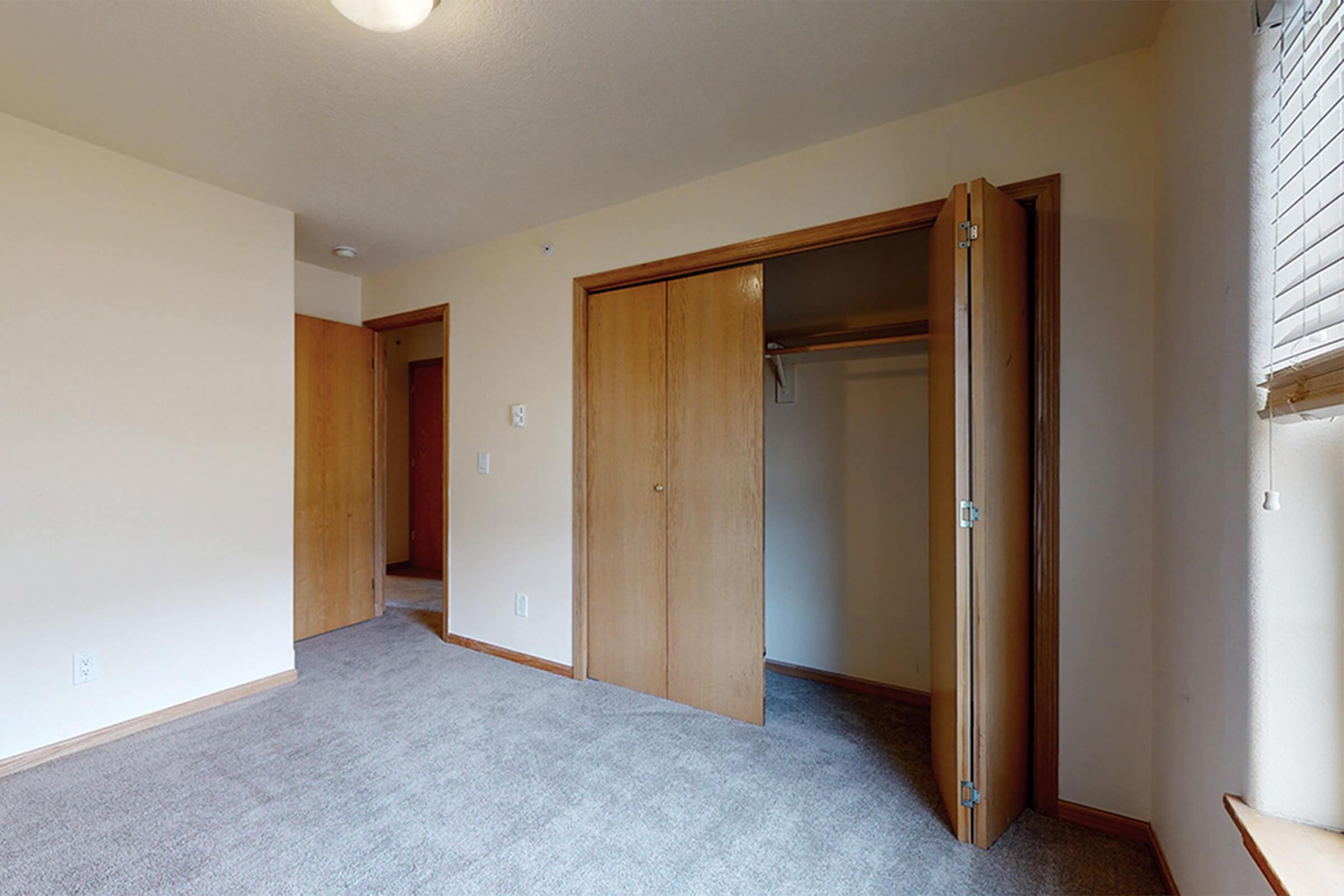 A spacious room featuring beige walls and gray carpet. There are two doors: one leads to a closet with sliding doors, and the other opens to another room. Natural light filters in through a window, illuminating the neutral-toned interior. The overall atmosphere is clean and inviting, suitable for various purposes.