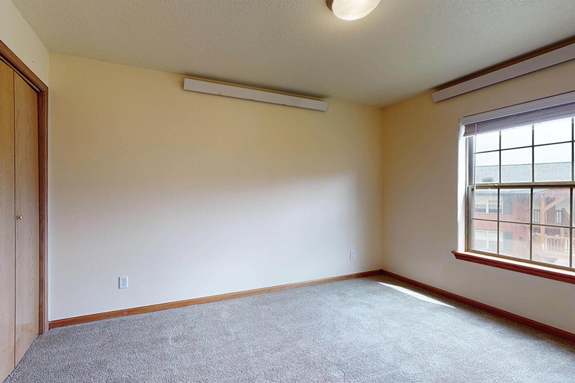 A vacant room with light beige walls and a large window allowing natural light. The floor is covered in gray carpet. There is a simple ceiling light fixture and a wooden door on one side, giving the space a clean and minimalist appearance.