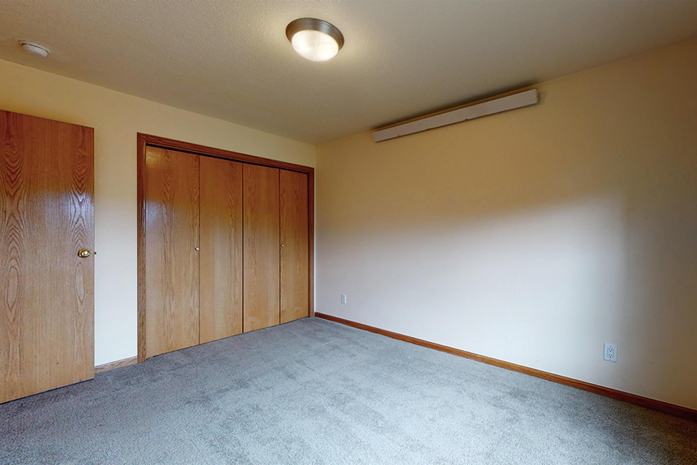 A simple, empty room featuring light-colored walls, a carpeted floor, and a ceiling light fixture. There are two wooden doors on the left leading to closets, while the right wall is mostly bare with a strip of light. The space appears clean and well-lit, ideal for various uses.