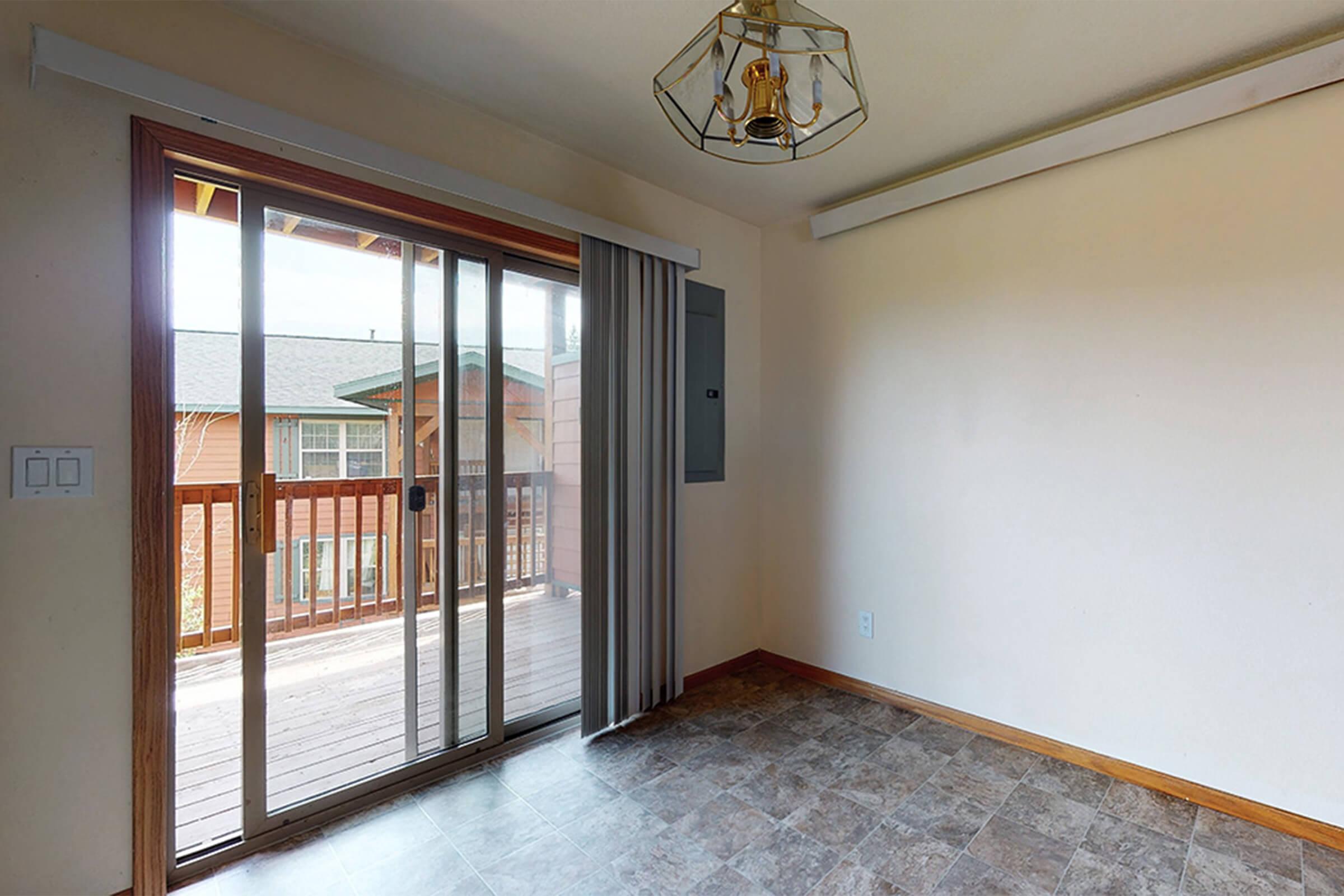 A spacious, empty room featuring a sliding glass door that opens to a wooden deck. The room has a light fixture hanging from the ceiling and a textured floor. Light enters from outside, illuminating the neutral-colored walls and the doorway leading to the deck area.