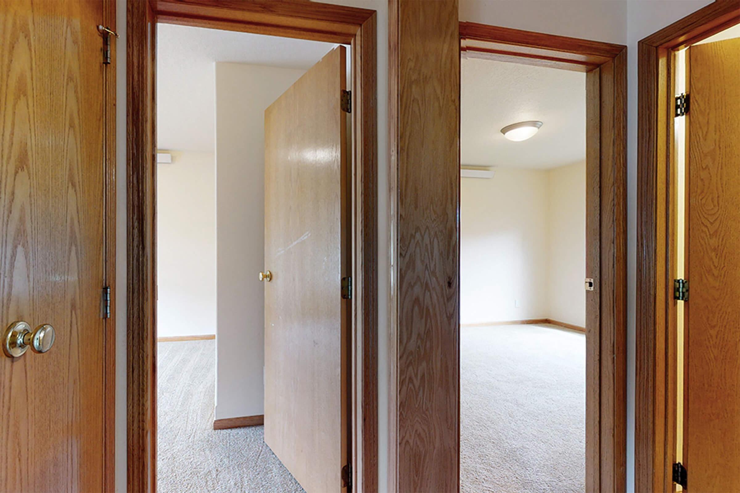 A view from a hallway featuring three wooden doors leading to empty rooms, with beige walls and carpeted flooring. Soft lighting comes from a ceiling fixture, creating a warm atmosphere. The space appears clean and well-maintained, ready for occupancy.