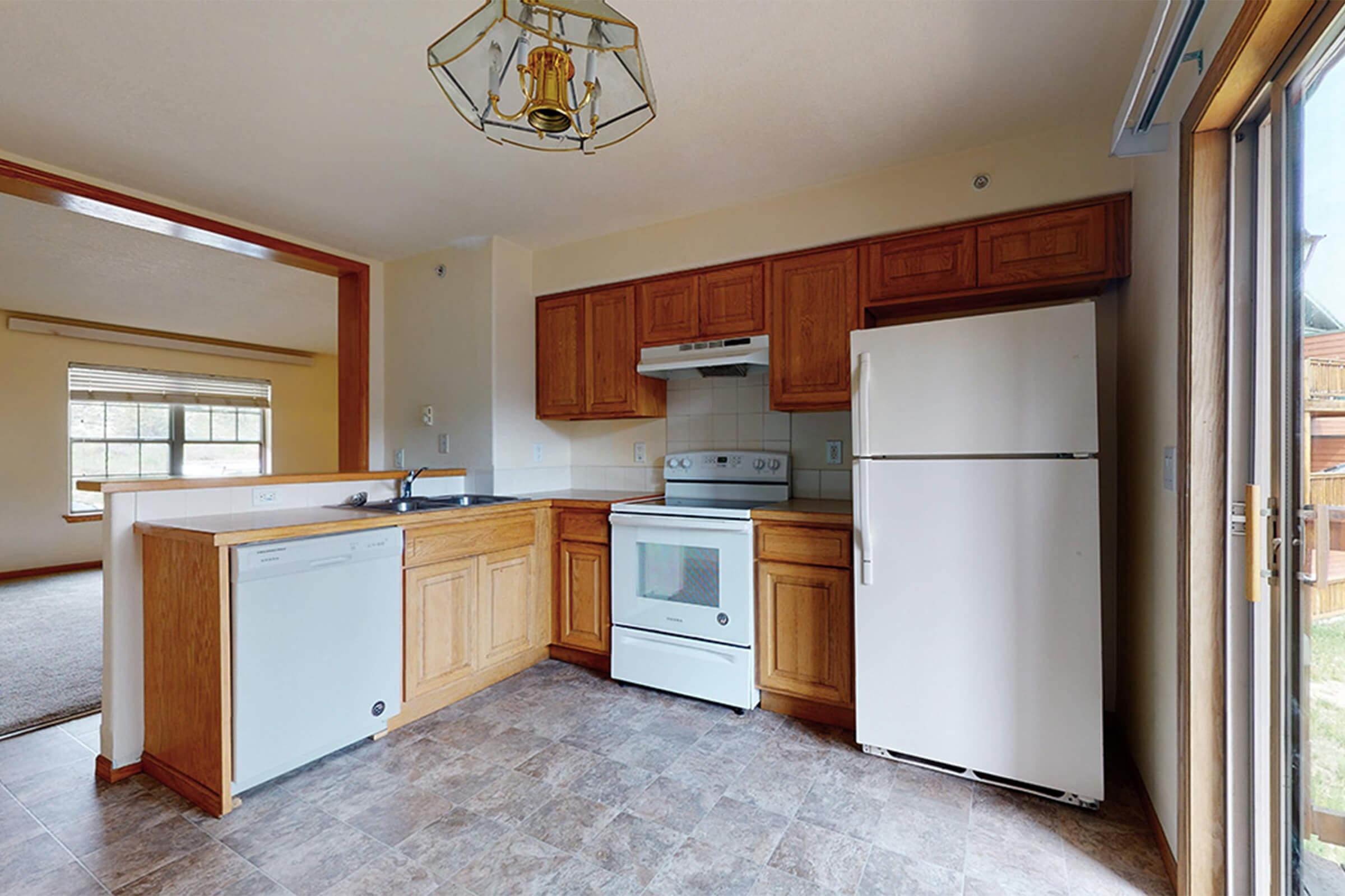 A kitchen featuring wooden cabinetry, a white stove, and an upright refrigerator. It includes a dishwasher, a sink, and a small countertop area. Natural light enters through a large window, and the flooring is a patterned tile. The adjacent space is visible, suggesting an open layout.