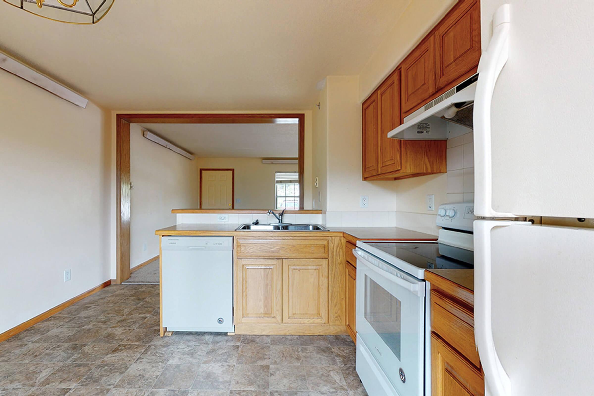 A kitchen area featuring beige walls, wooden cabinetry, a white refrigerator, an oven, and a dishwasher. The floor is tiled in a light gray pattern. A window provides natural light, and a light fixture hangs from the ceiling. The space has an open layout, connecting to a living area.