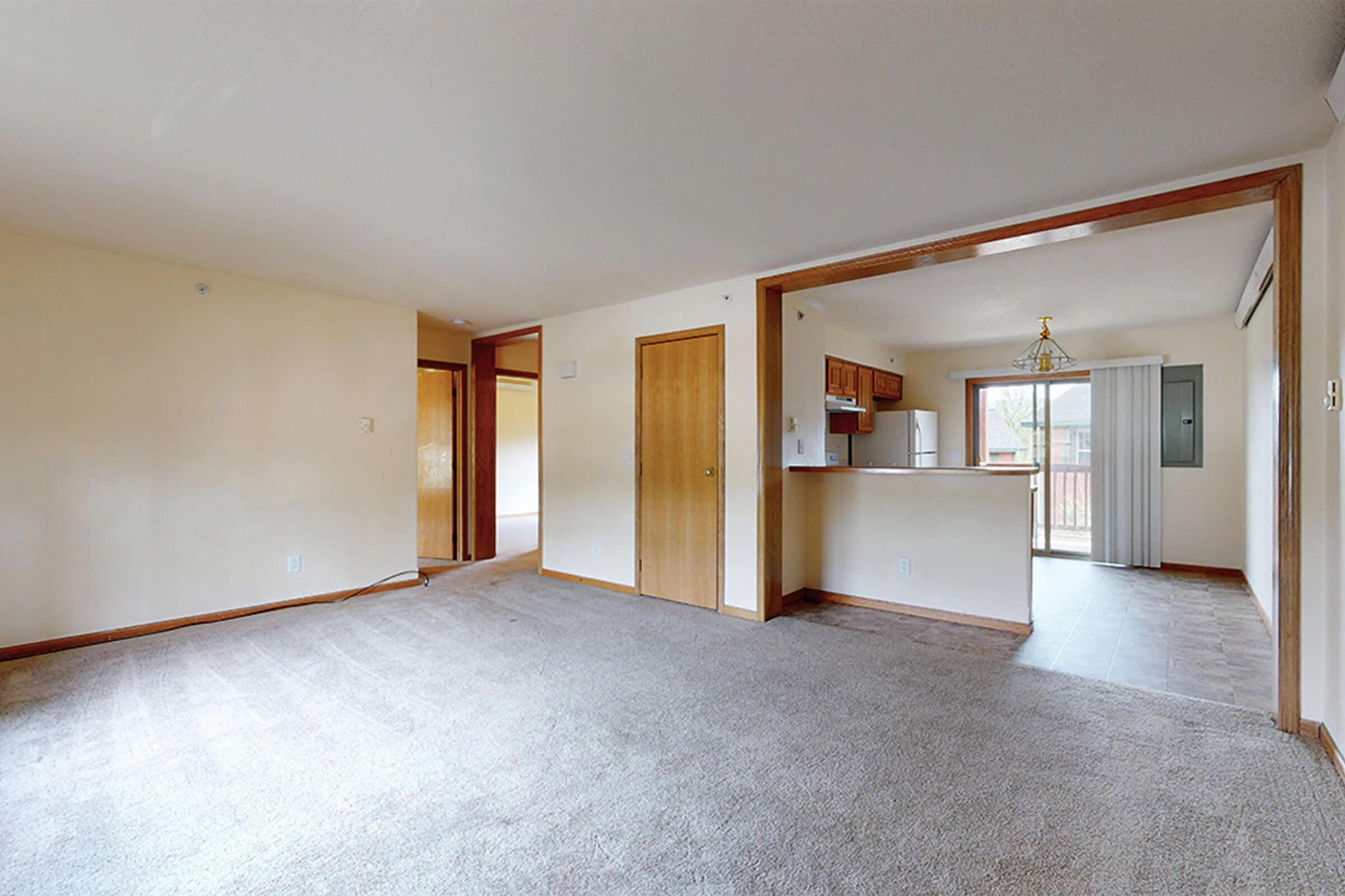 A spacious, well-lit living room with beige walls and carpeting, leading to a kitchen area. The kitchen features wooden cabinetry and a window, with a dining area visible. The overall space is empty, suggesting it is ready for new occupants.