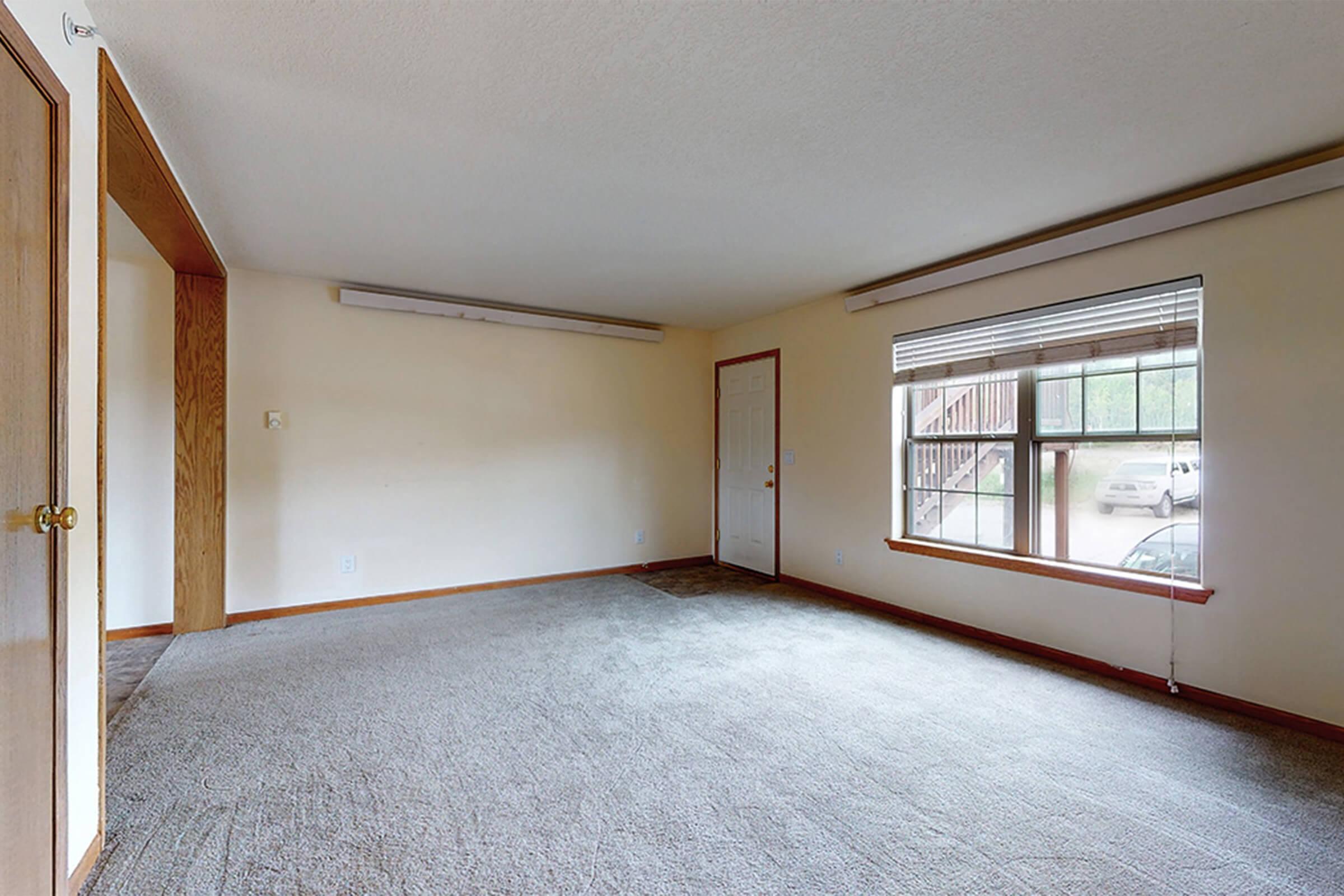 A spacious, empty room with beige carpeting and light-colored walls. There is a large window with blinds letting in natural light, a door leading outside, and an opening to another room on the left. The overall atmosphere is bright and airy, ideal for various design possibilities.