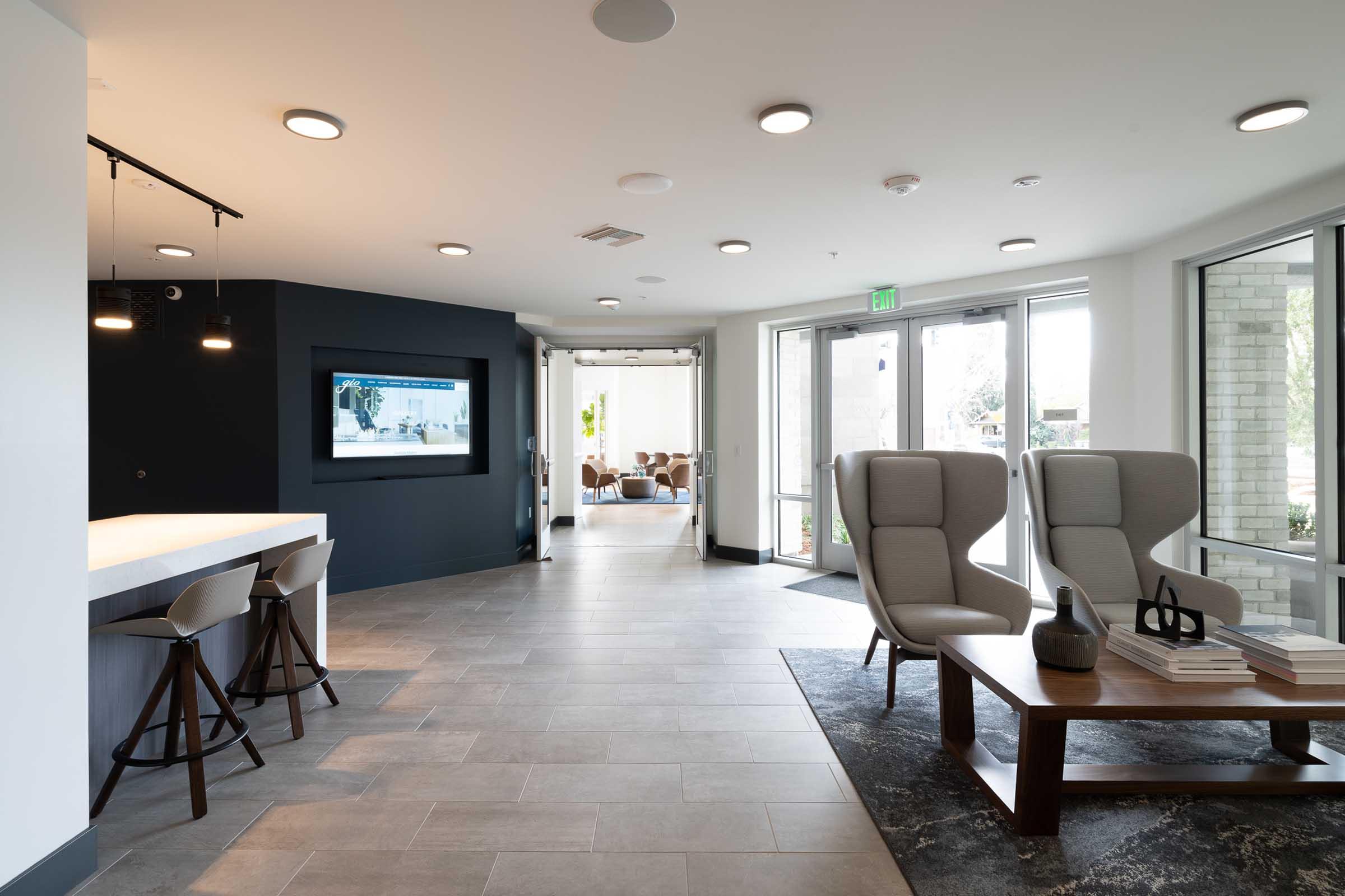 Modern lobby interior featuring a light color palette, a sleek bar area with stools, and comfortable seating. Large windows allow natural light. A modern television mounted on a dark wall adds a contemporary touch, while a stylish coffee table and decorative items enhance the inviting atmosphere.