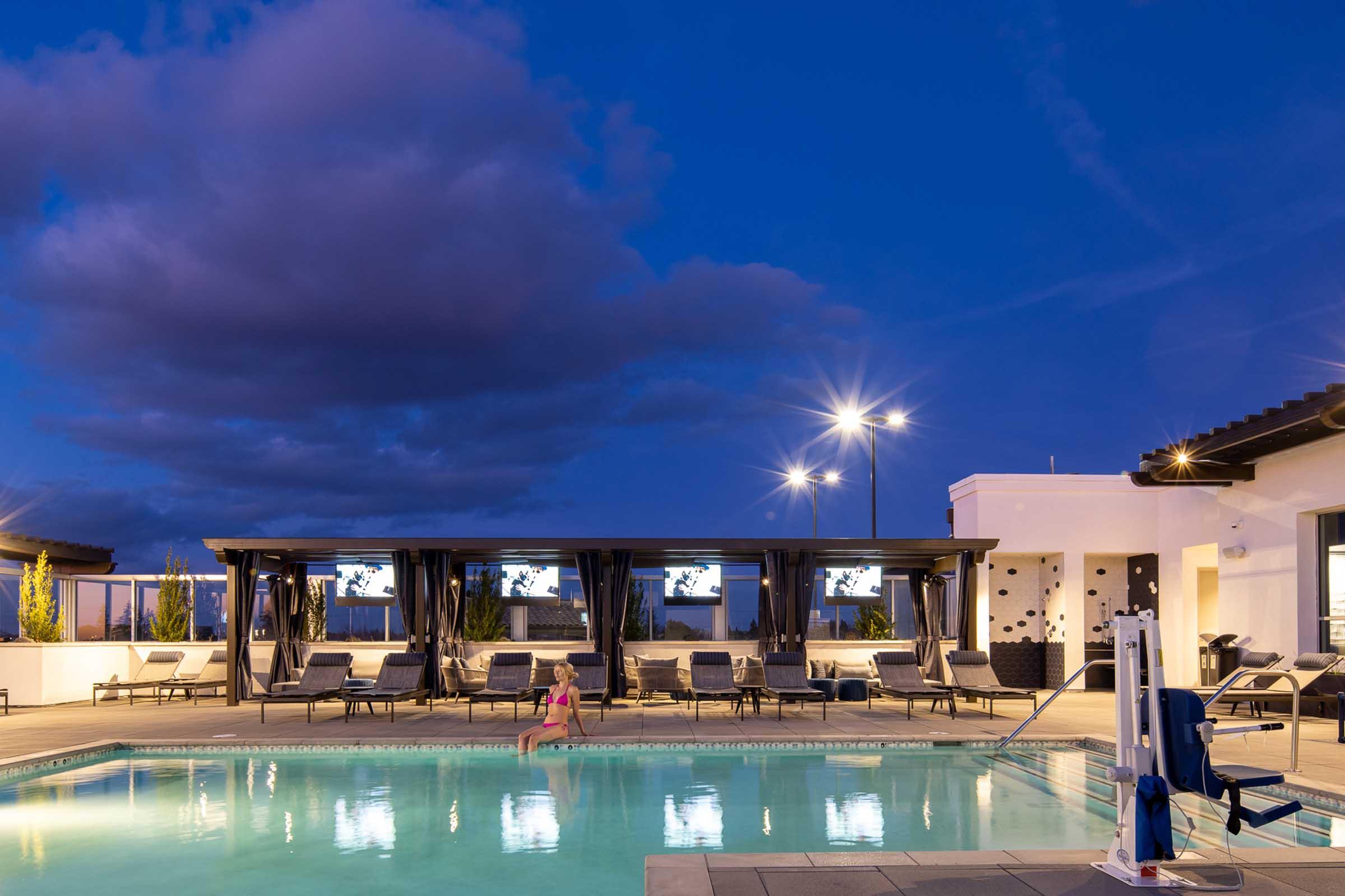 A rooftop pool area at dusk, featuring a well-lit swimming pool surrounded by lounge chairs. Several cabanas with curtains provide shade, and large screens are visible in the background. Soft clouds scatter across the evening sky, adding to the tranquil atmosphere.
