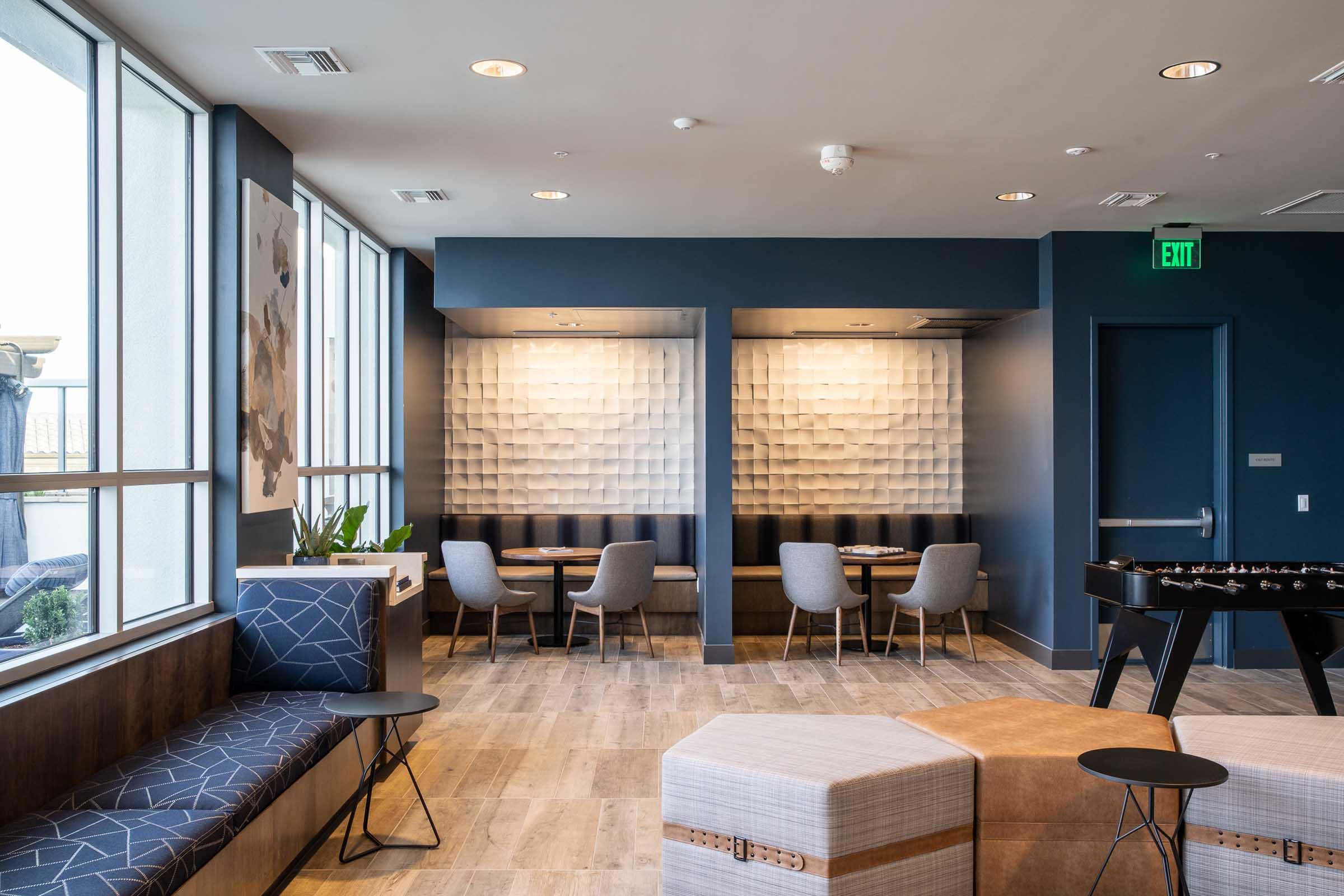 A modern lounge area featuring two distinct seating sections with grey chairs and stylish tables. The walls are adorned with textured panels, and there are decorative elements like plants. Natural light filters in through large windows, creating a welcoming and contemporary atmosphere.