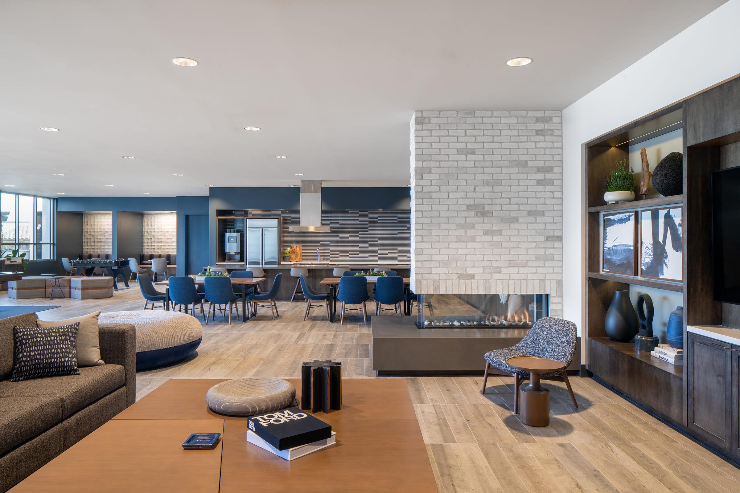 A modern, stylish interior of a spacious lounge area featuring a sectional sofa, a coffee table, and a unique chair. The room includes a fireplace, decorative shelving, and a large window with plenty of natural light. Dining tables and chairs are visible in the background, creating a welcoming atmosphere.