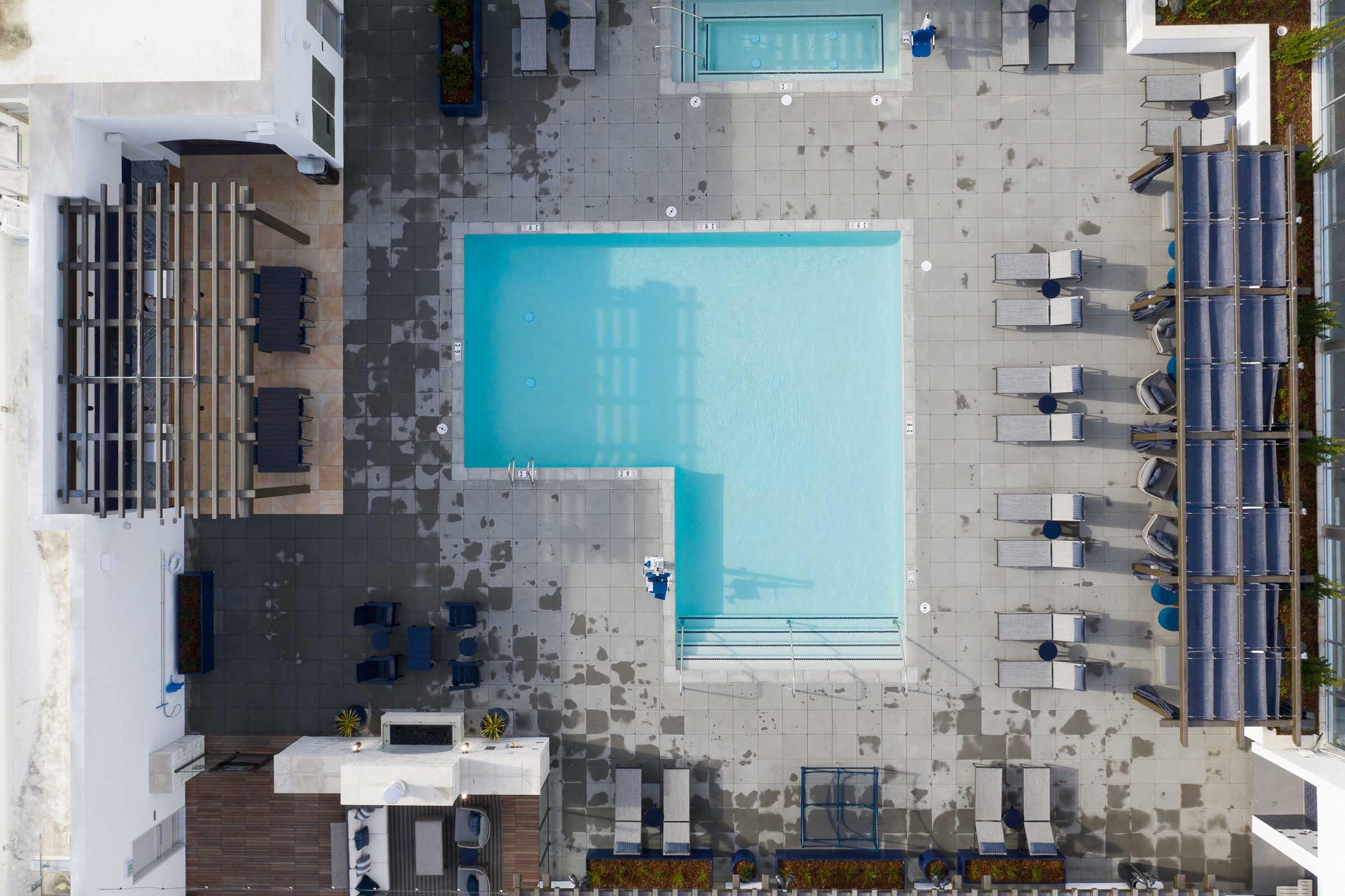 Aerial view of a modern rooftop pool area featuring a rectangular swimming pool surrounded by gray lounge chairs, umbrellas, and planters. The pool is clear blue, with a few poolside chairs arranged neatly. Wet floor surfaces suggest recent cleaning or rain.