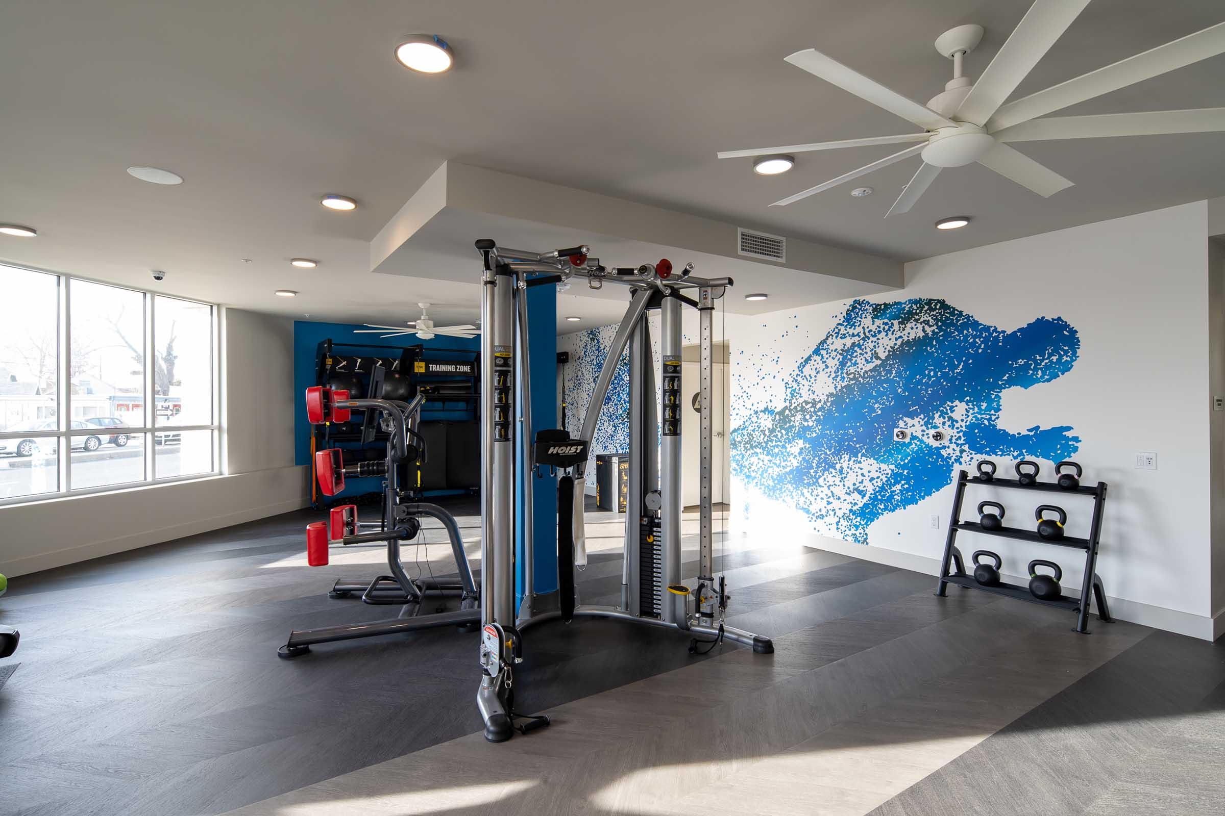 A modern fitness room featuring various workout equipment, including a multi-functional weight machine, free weights, and kettlebells. Large windows provide natural light, and a blue abstract mural decorates one wall, enhancing the contemporary aesthetic. Ceiling fans help with ventilation.